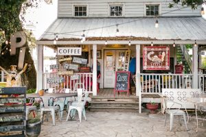 A small, white farmhouse-style building with several signs that read words like "Pie Haven" and "Coffee." There is a cobblestone porch with tables for patrons.