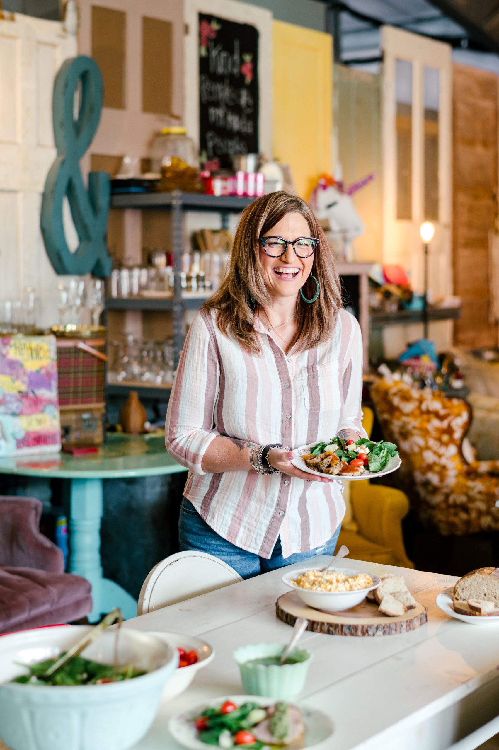 A smiling woman in a striped shirt and glasses holds a plate of salad inside a kitschy farmhouse shop