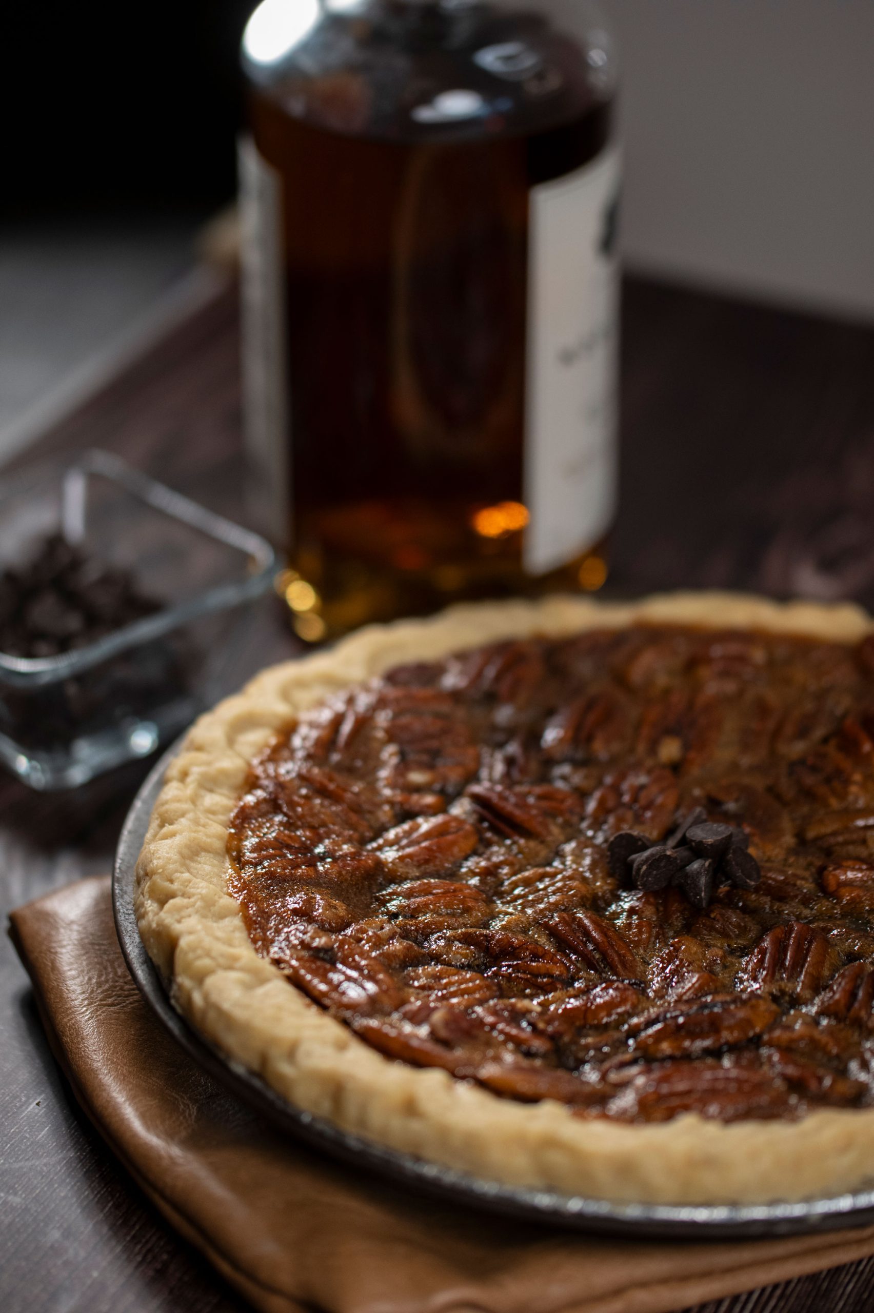 A pie with a pecan filling topped with chocolate bits beside a container of chocolate chips and a bottle of bourbon
