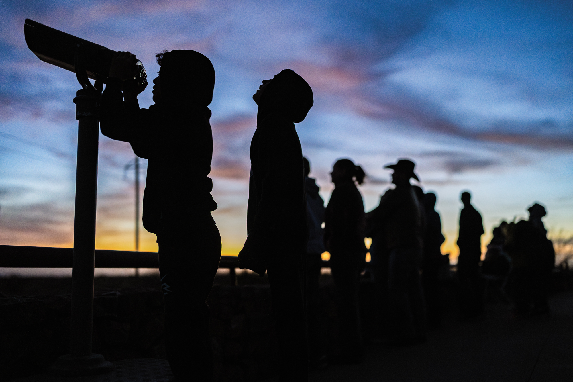Silhouettes of several people lined up on a pier watching the sunset. A little boy peers out a public viewfinder