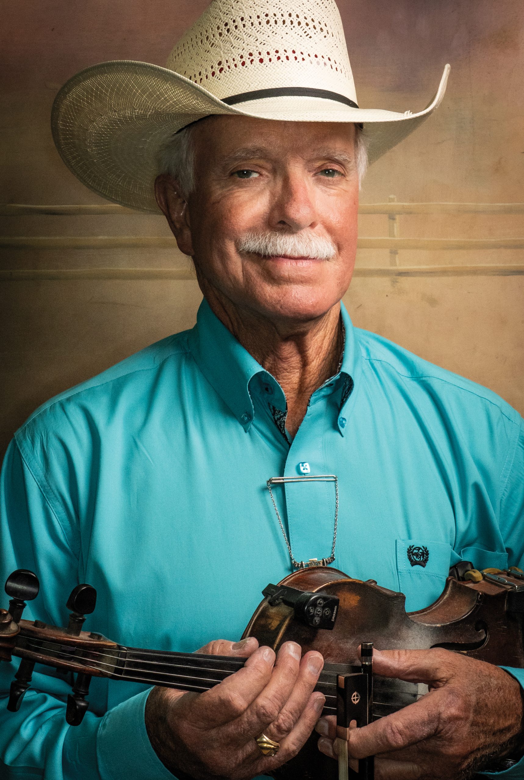 An older man in a teal button down shirt and an ivory cowboy hat holds a fiddle