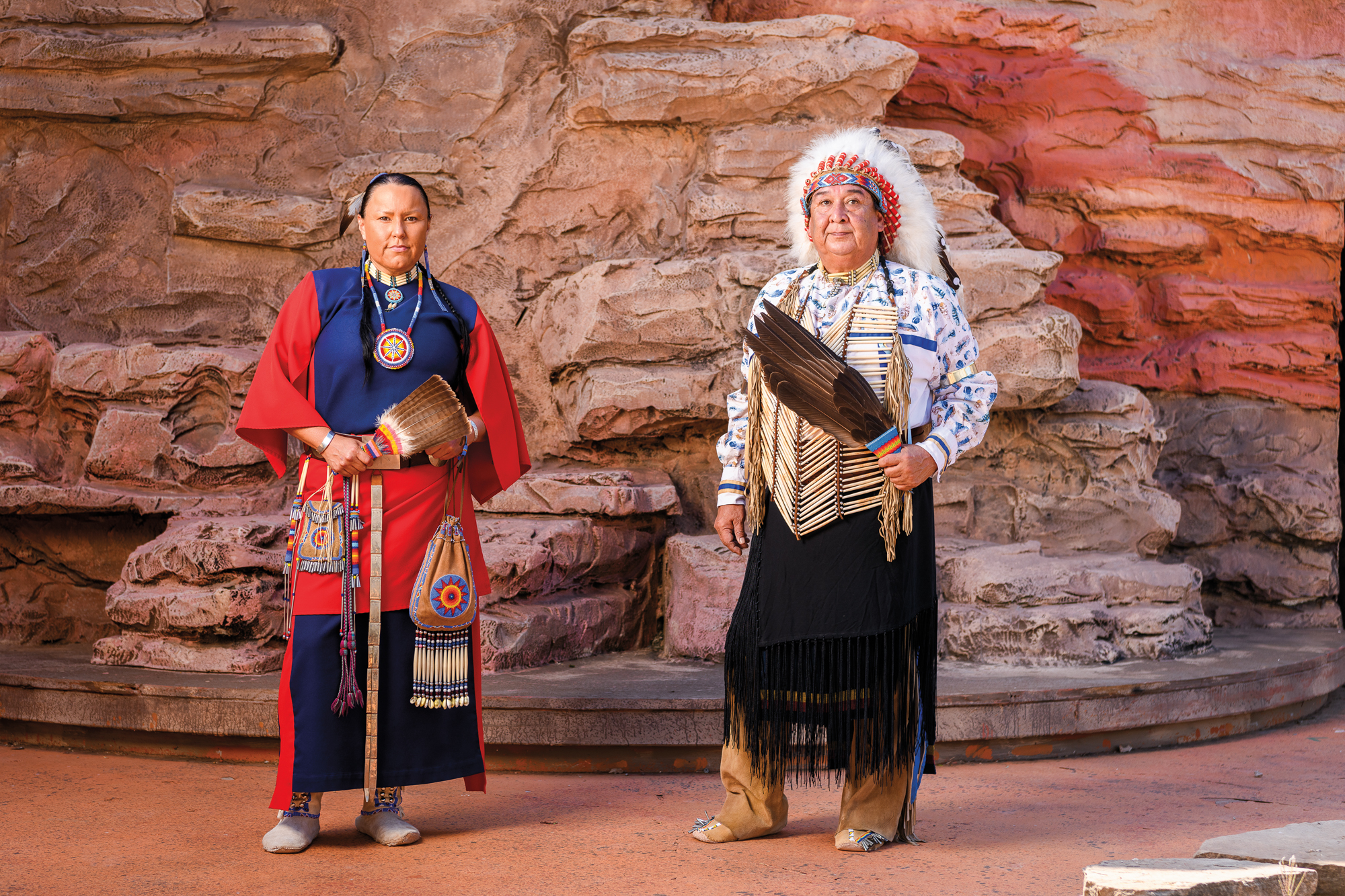 A man and woman in traditional Native American garb pose in front of red stone