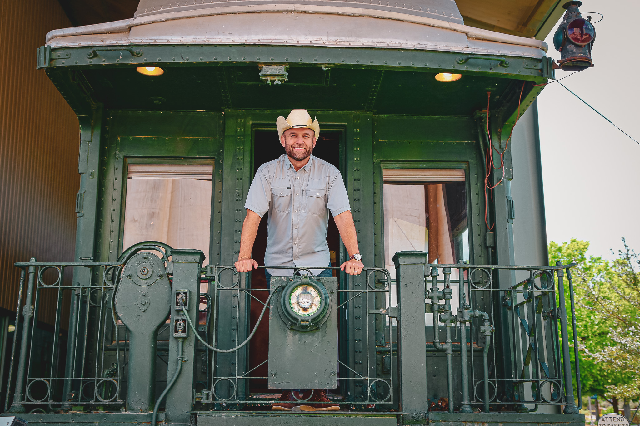 A man in a button down and cowboy hat stands on the caboose of a green train