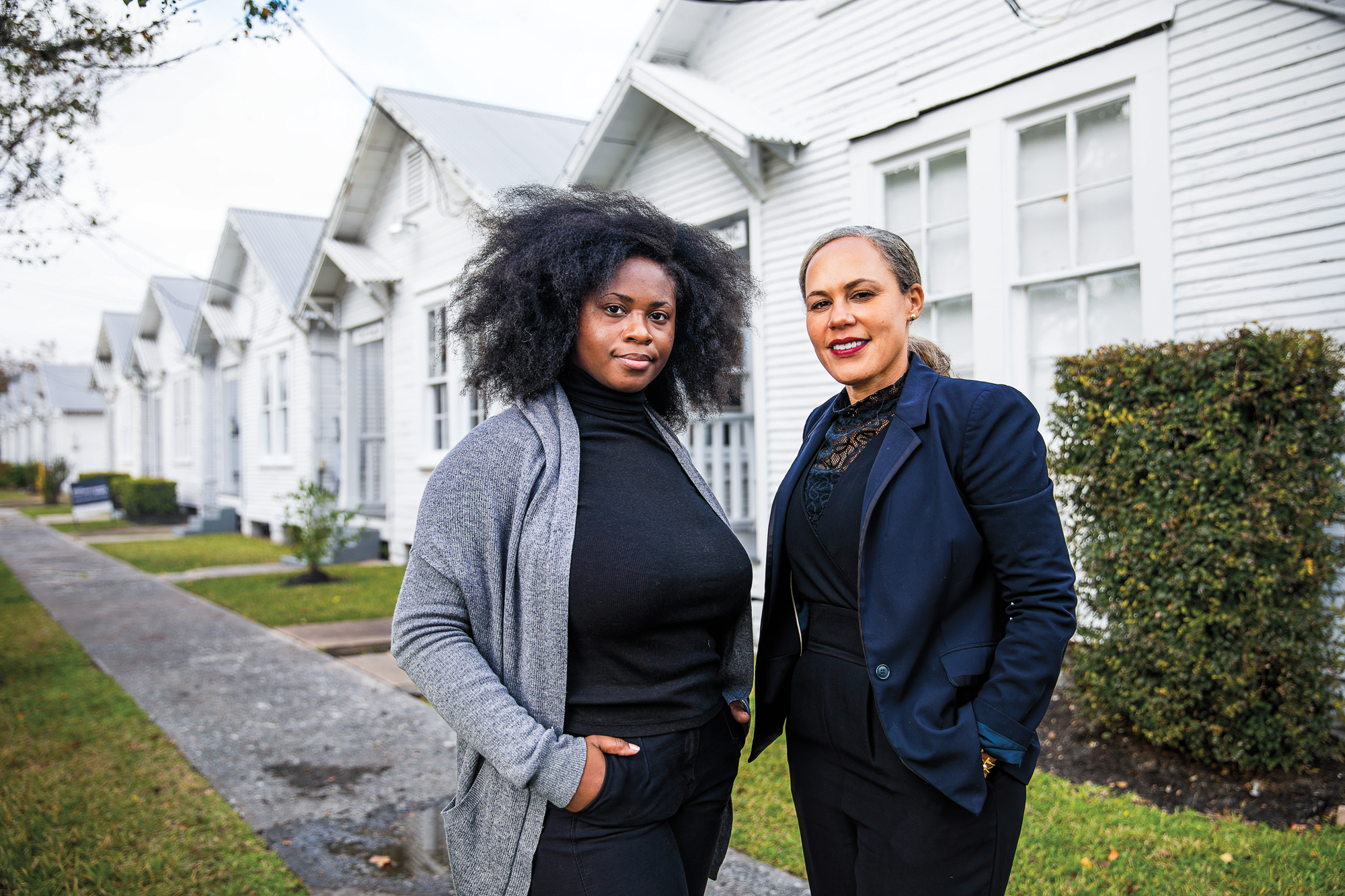 Two women in professional clothing pose in front of a row of white houses