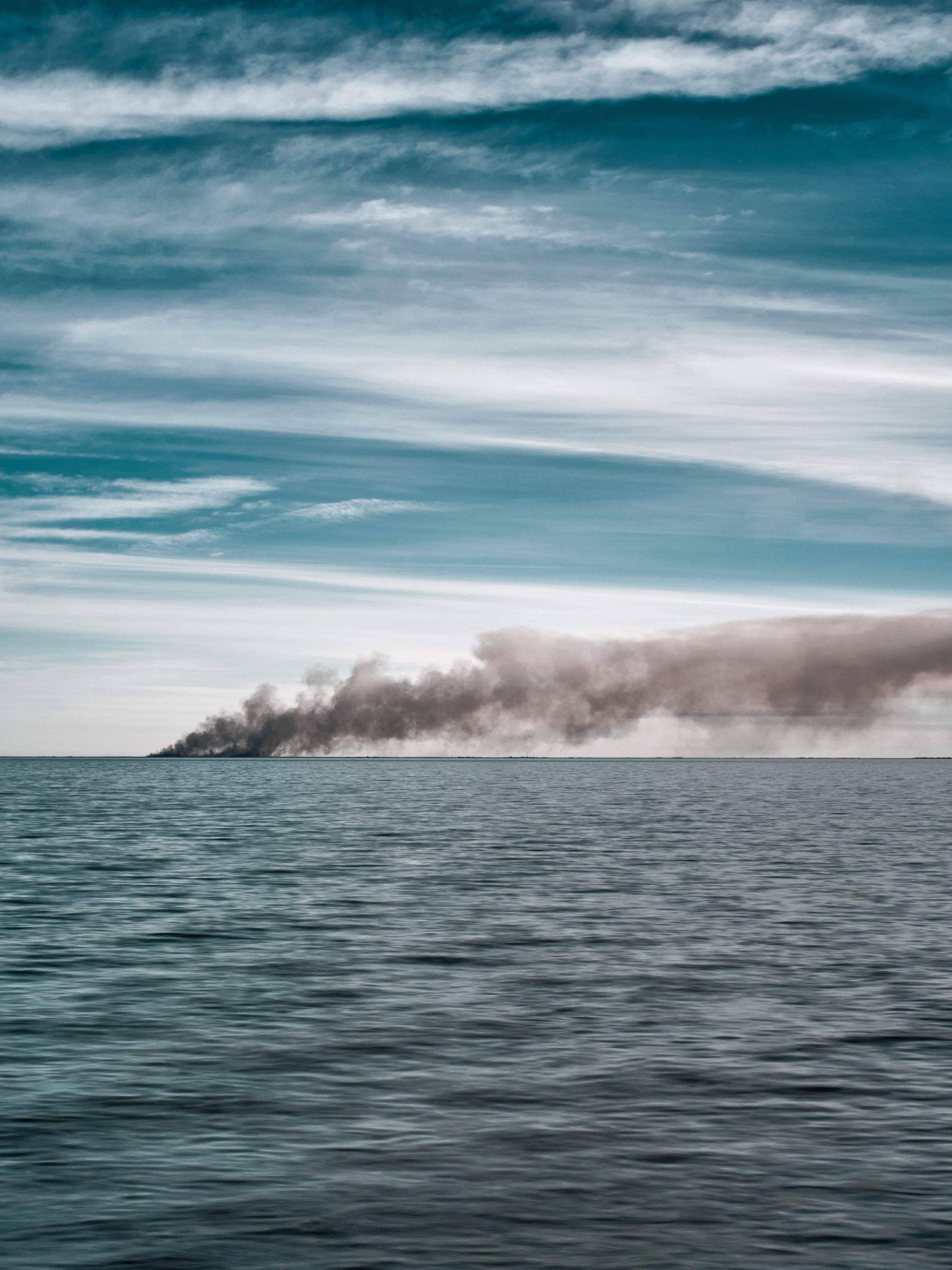 A large wave of dark smoke seeps into the sky on the ocean horizon