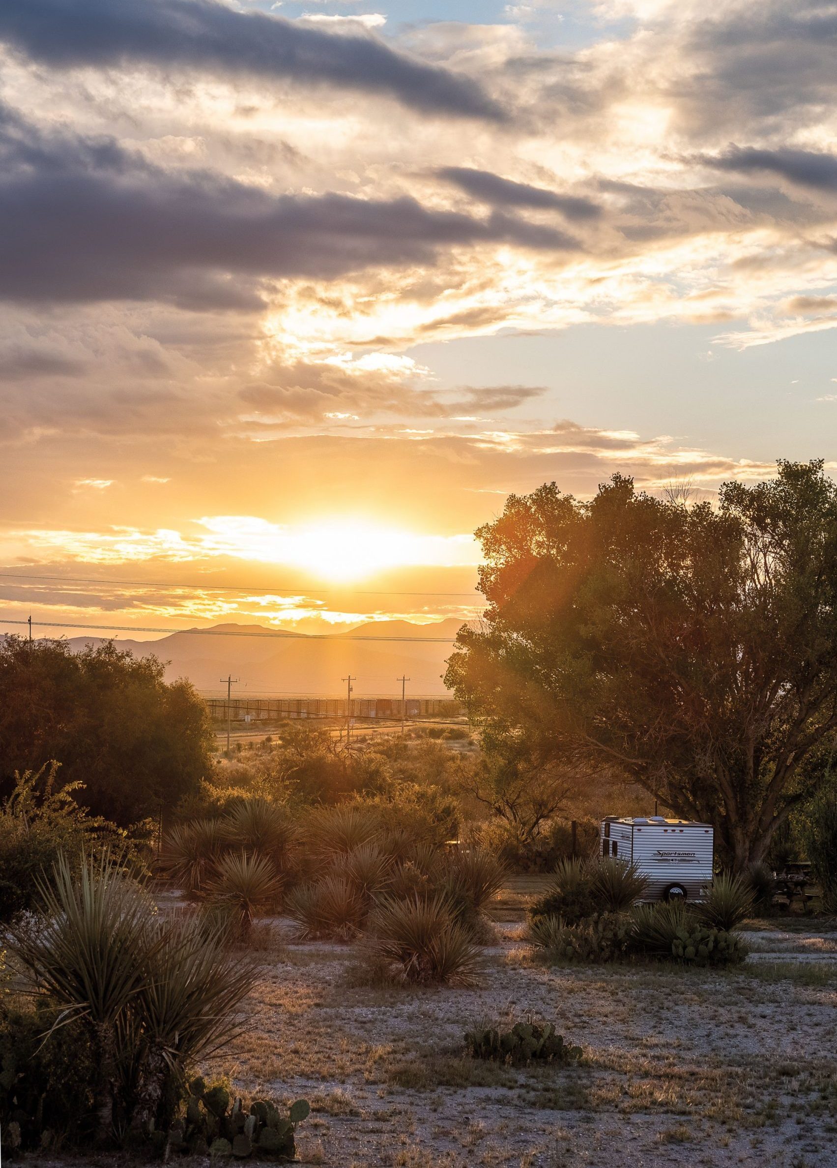 An RV camper is parked among trees surrounded by a mountainous desert landscape at sunset