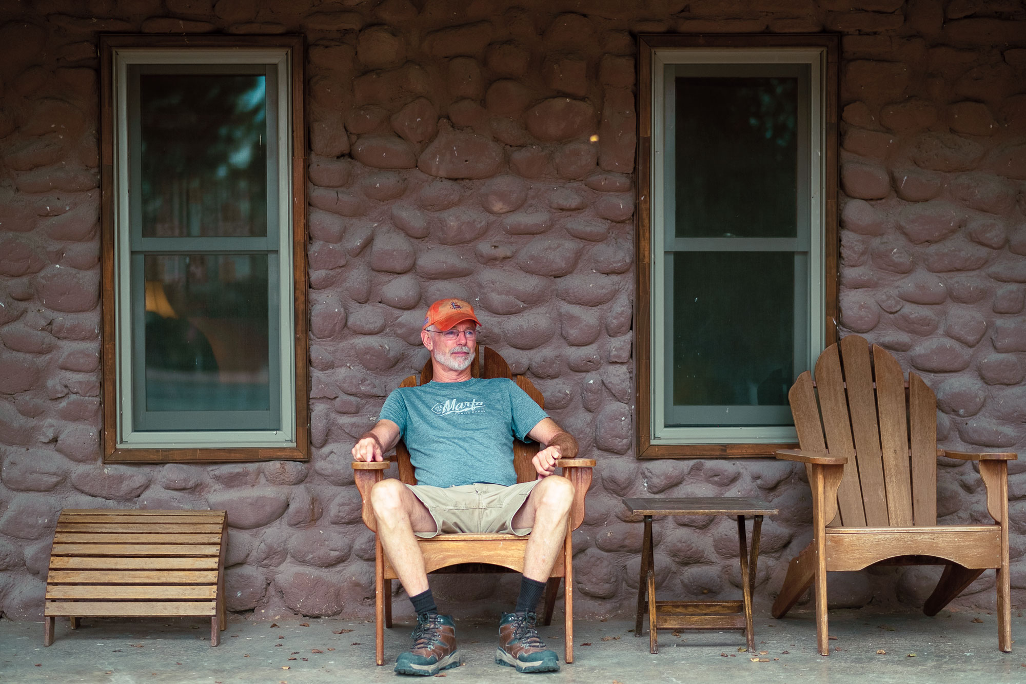 A man in a green shirt and red camp lounges on a chair outside a stone facade between two vertical windows