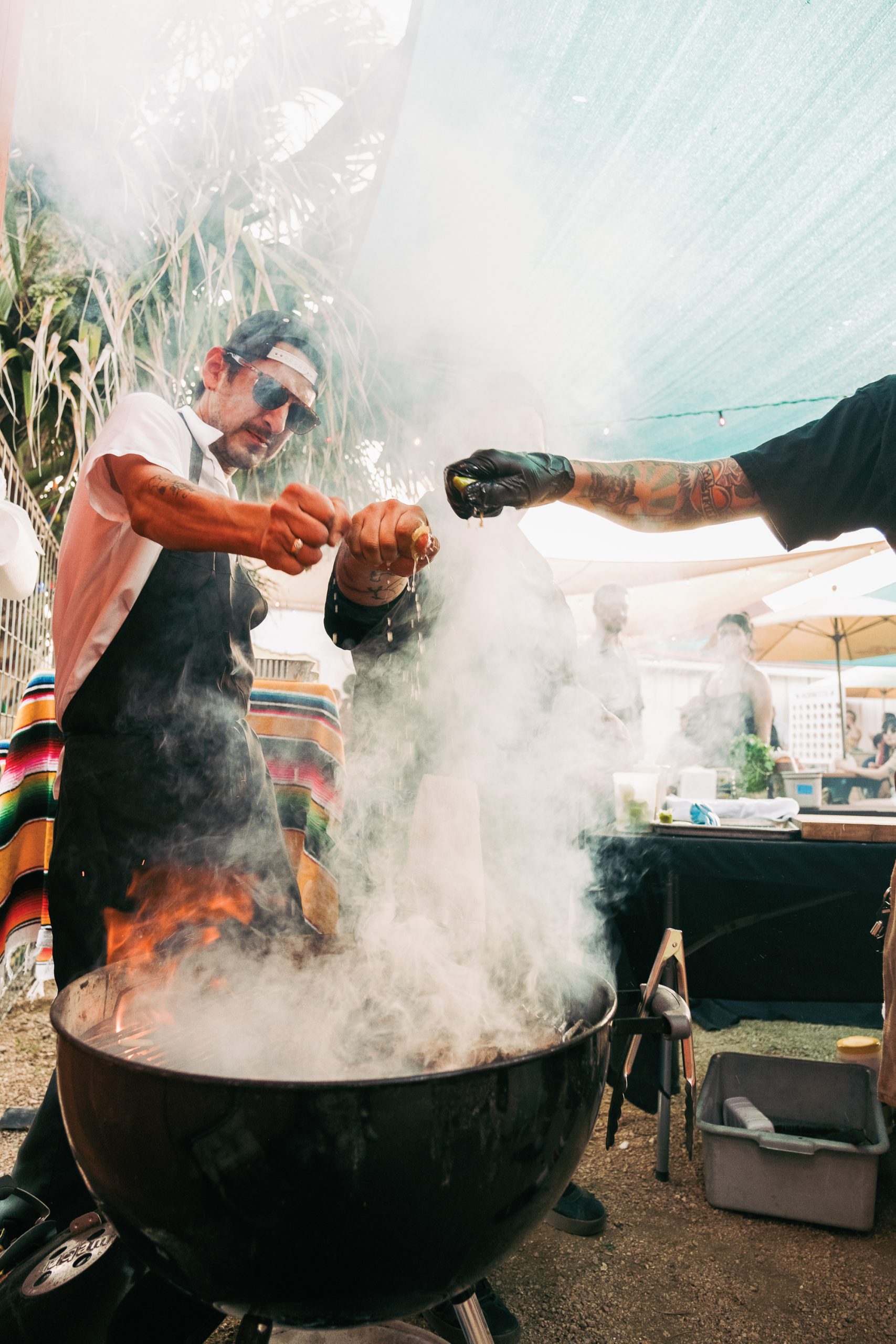 Three men in cooking attire squeeze limes over a sizzling grill