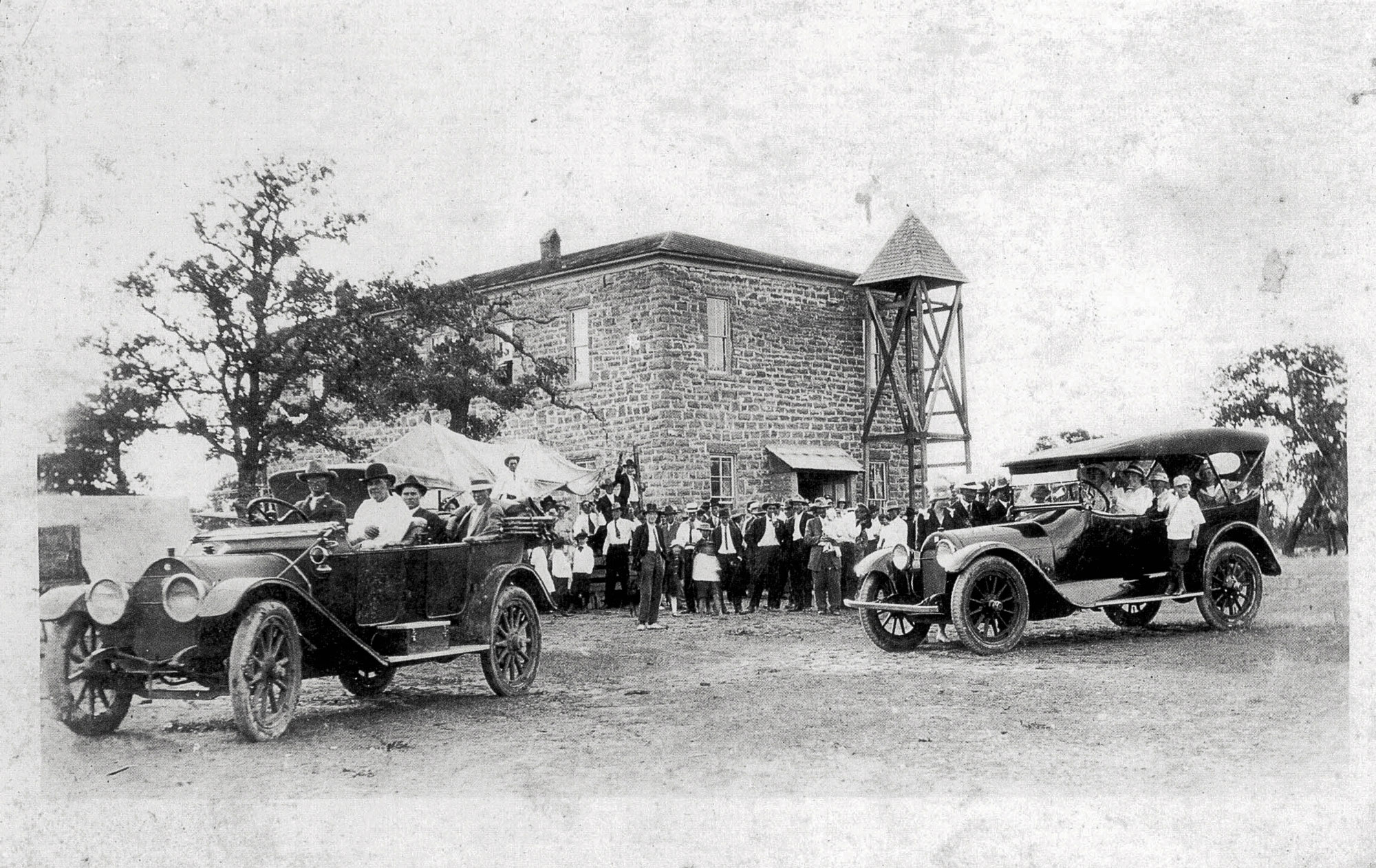 A black-and-white photograph of two cars and many people standing in front of an old schoolhouse