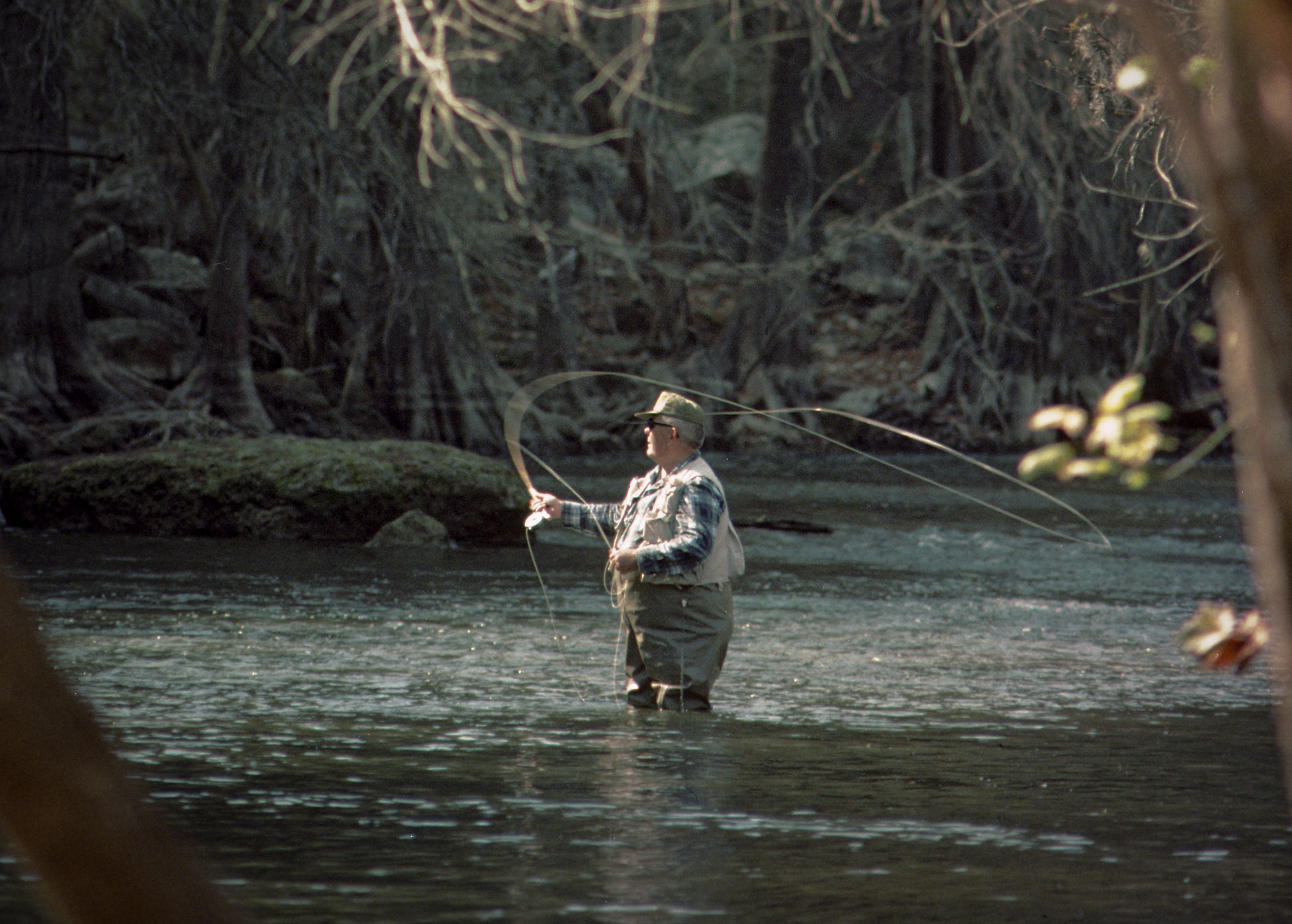 A man in glasses fly-fishing in a river with the water up to his knees