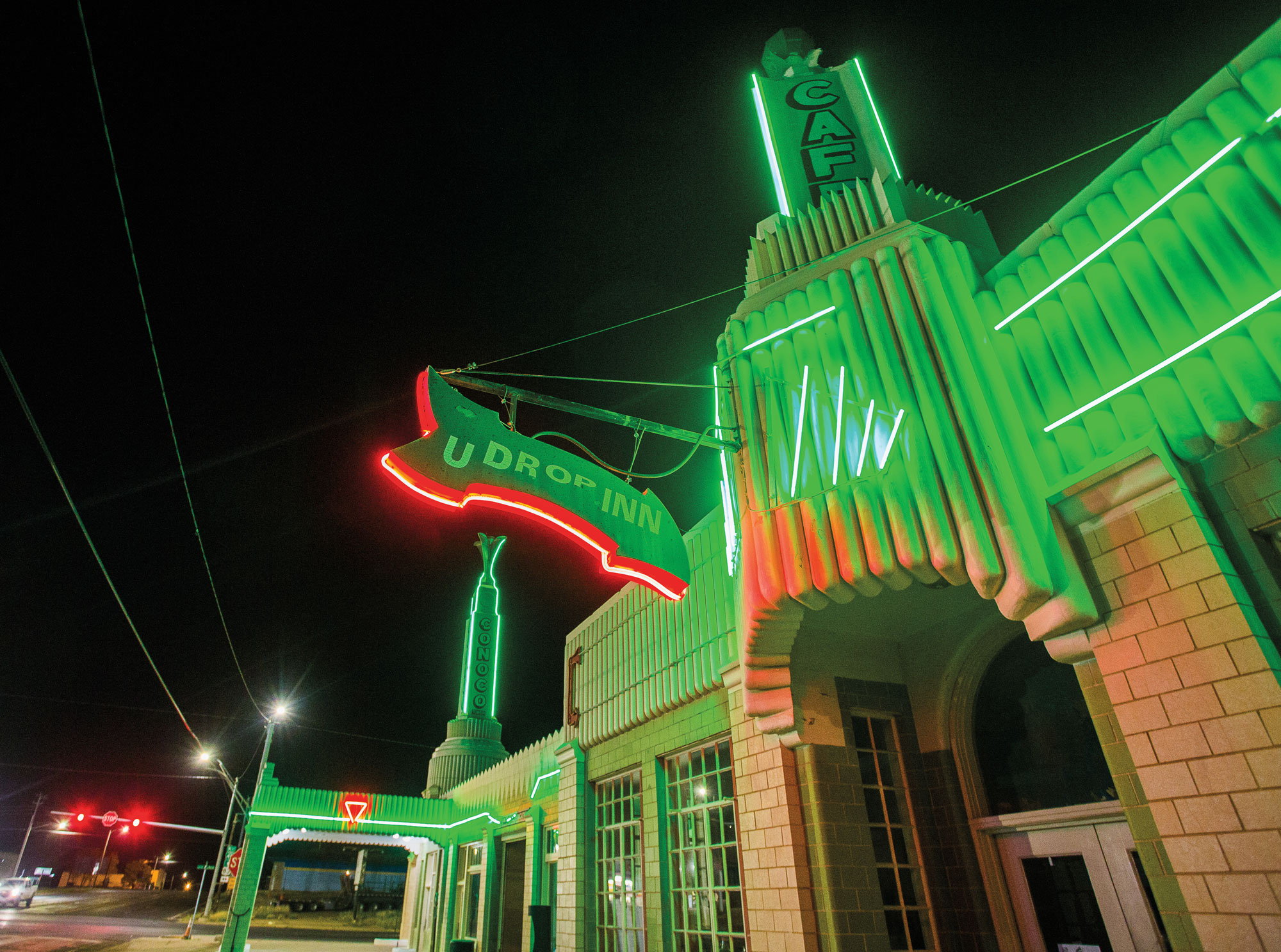 An art deco-style building with signs that read "U Drop Inn" and "Cafe" is lit a neon green at night.