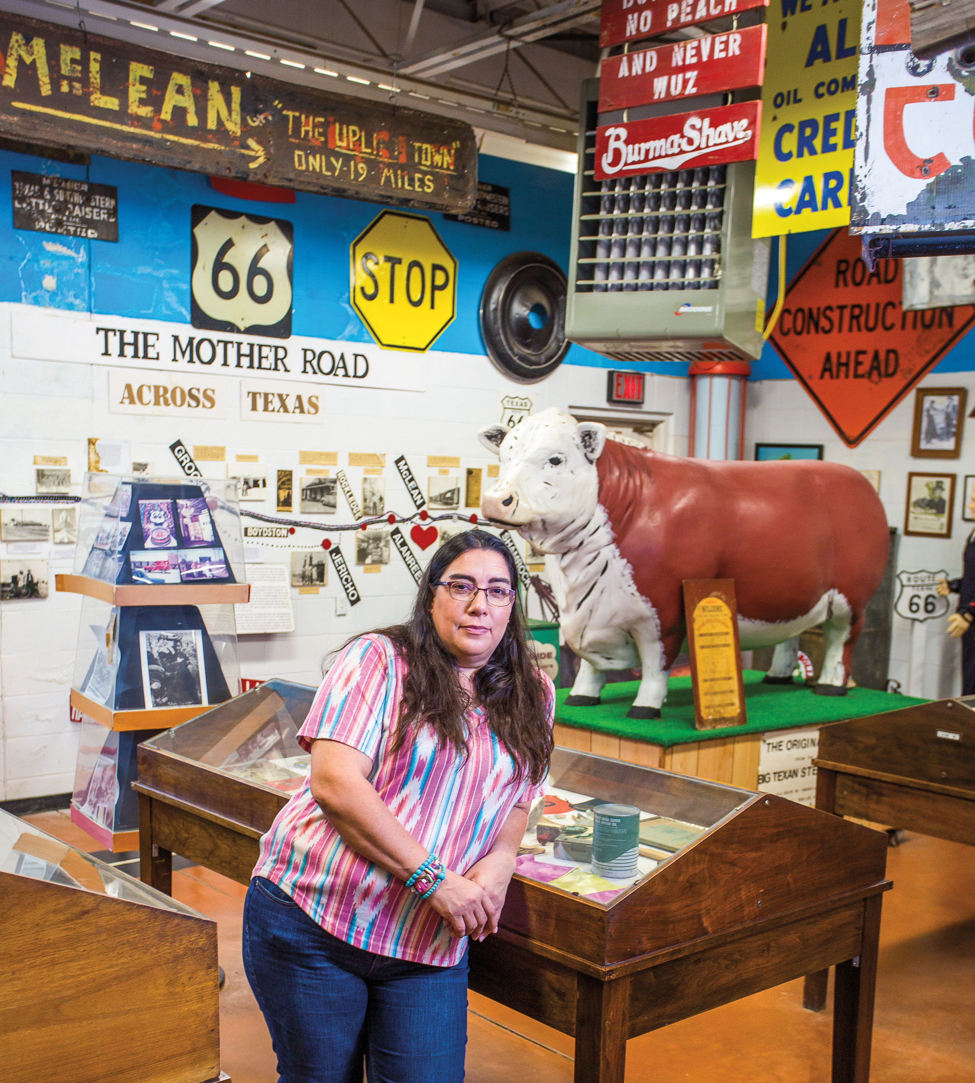 A woman leans on a glass display case in an eclectic room filled with road signs that read phrases like "The Mother Road" and "Stop."