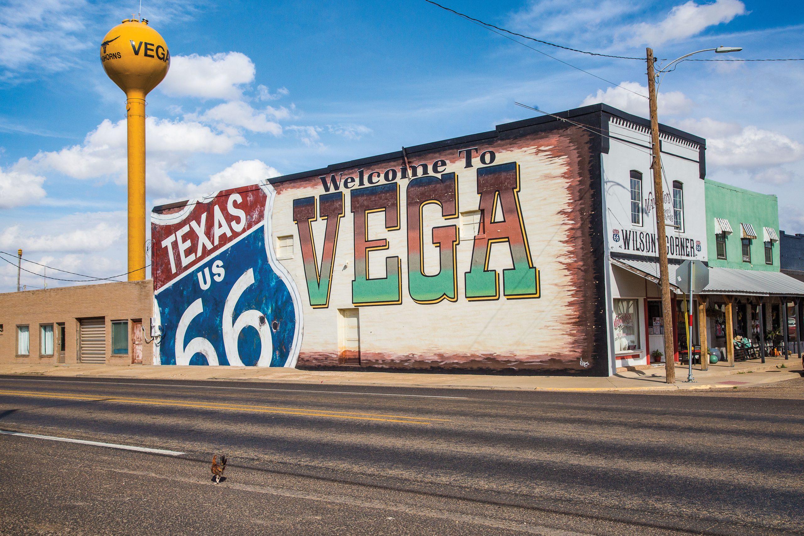 A yellow water tower and the side of a building with a mural of a sign that reads "Texas US 66" and "Welcome to Vega."