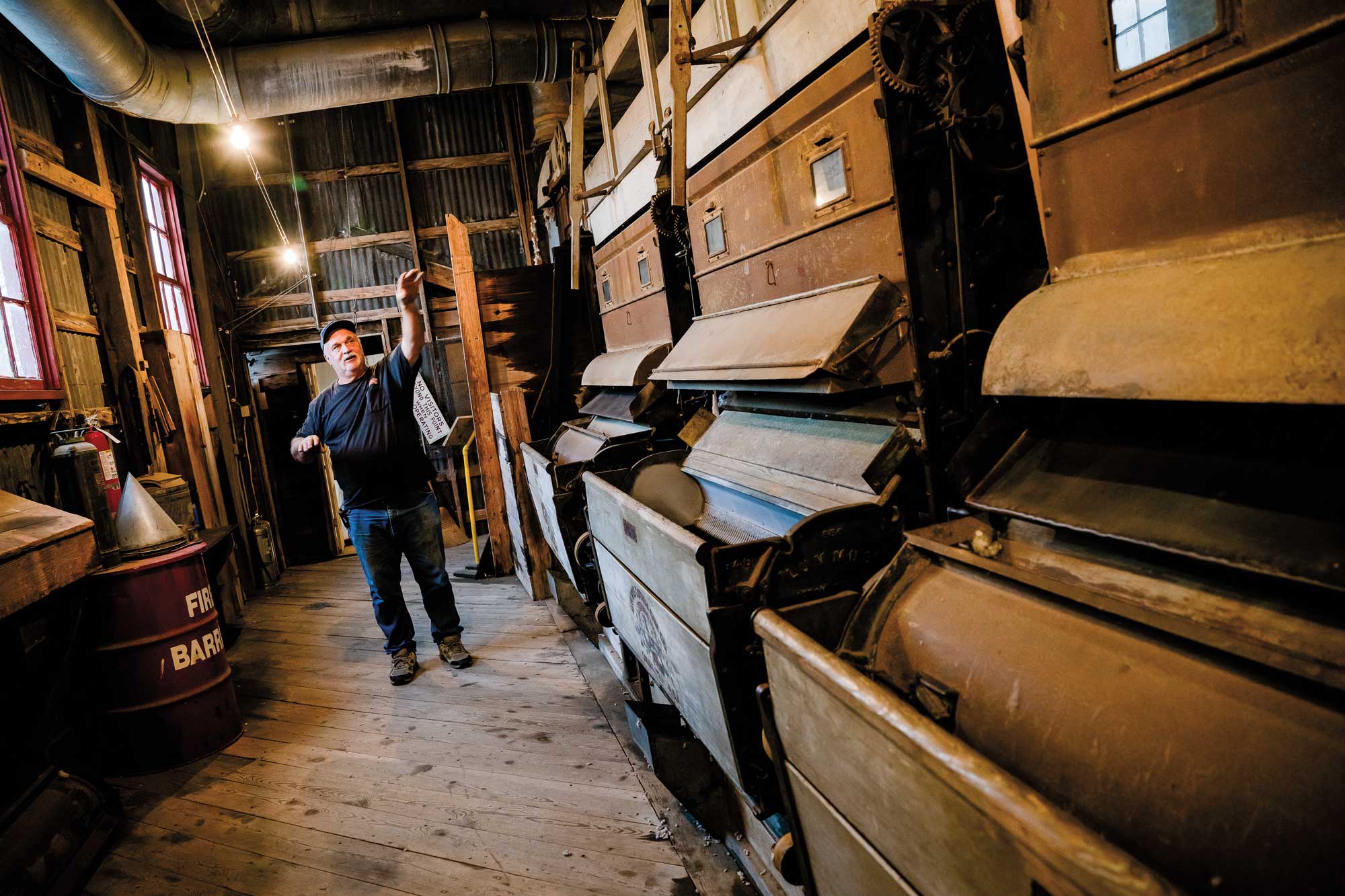 A man in a blue shirt and jeans pointing toward an old cotton gin