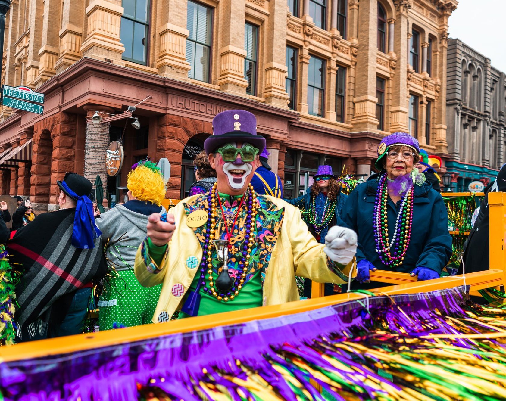 A man dressed in a purple, gold, and green clown outfit poses in a crowd