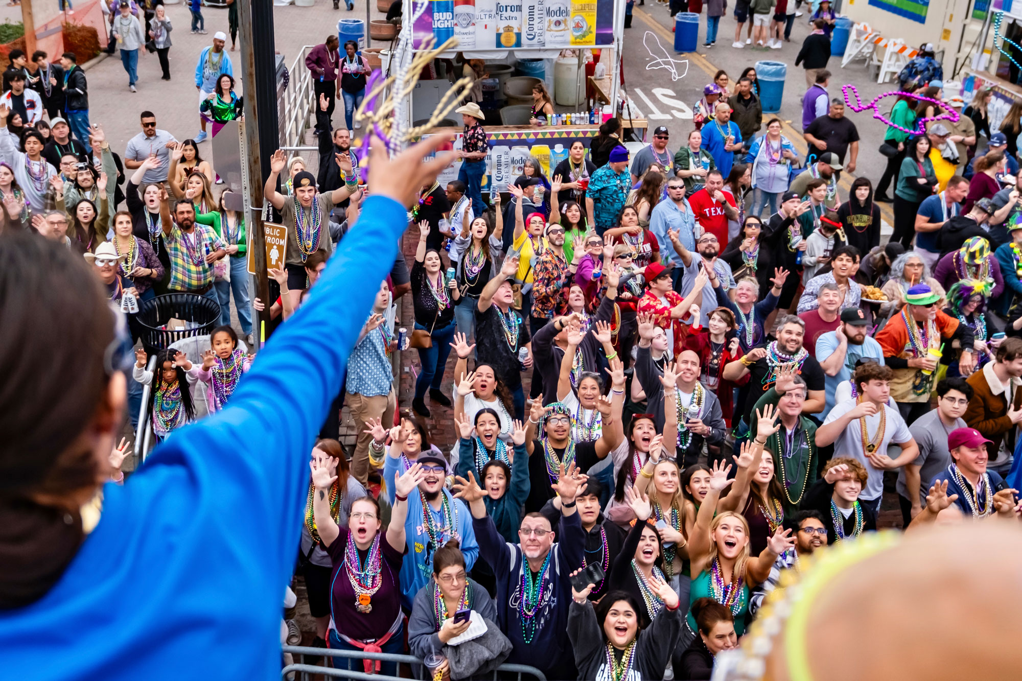 An arm reaches out from a balcony over a crowd of people dressed in purples, green, and yellow