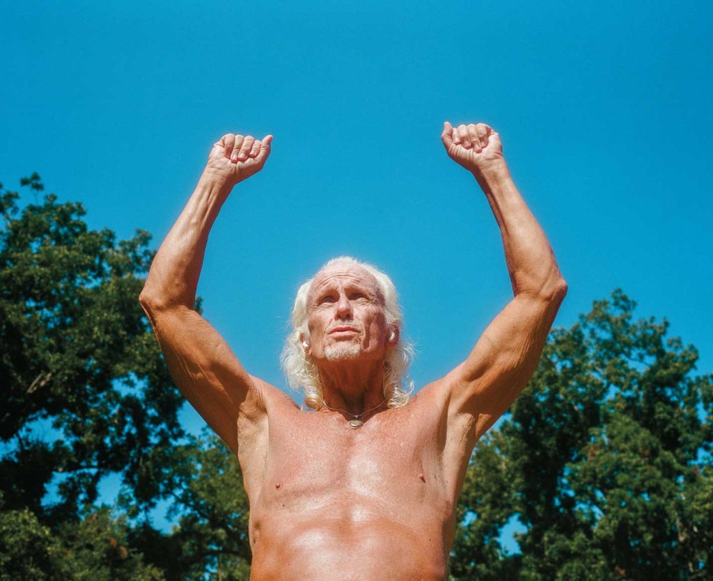 A tanned, fit, older man raises his arms over his head to the sky in a park