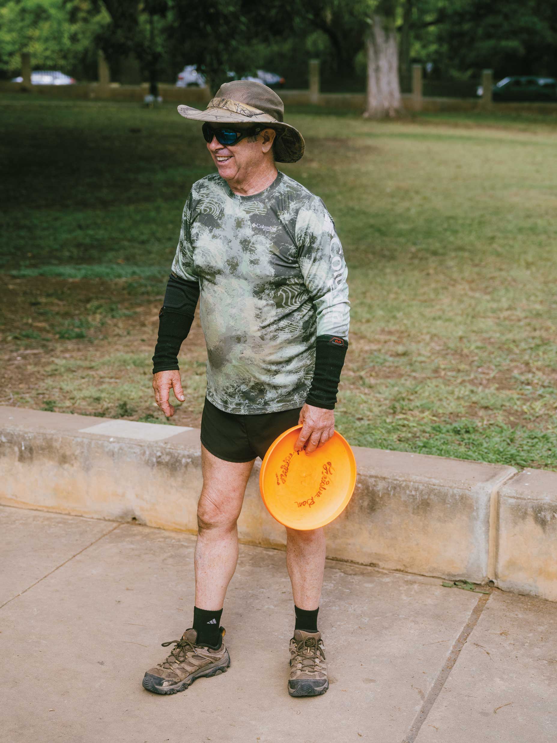 A man in a camo shirt, black shorts, sunglasses, and a hat holds a bright orange disc.