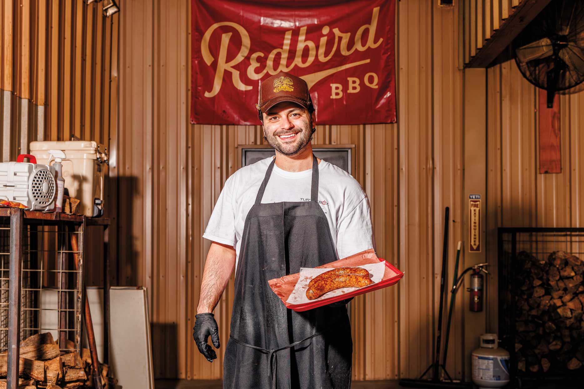 A man in a black apron, a red cap, and black gloves holds a tray of sausage under a flag that reads "Redbird BBQ."