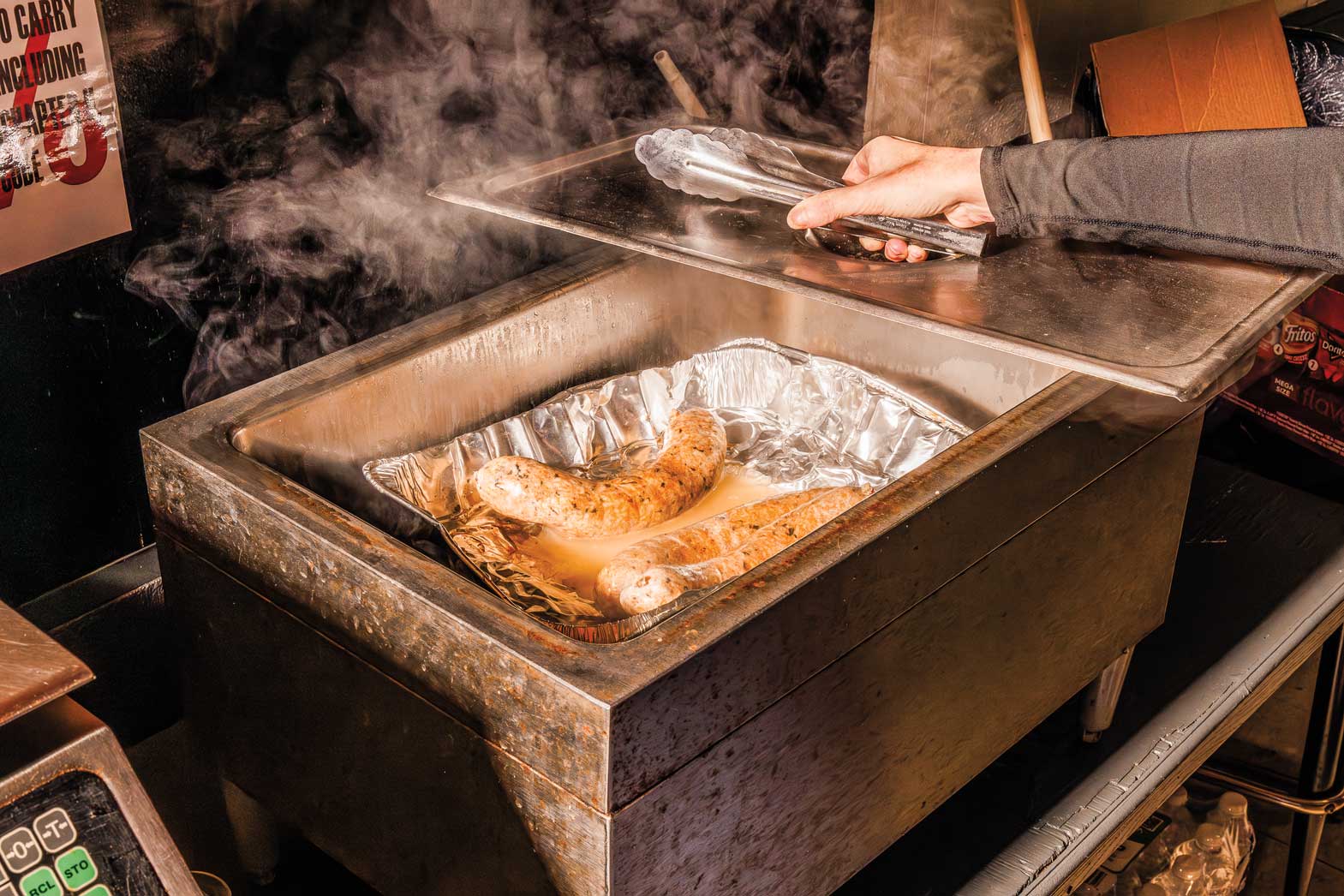A person holds tongs next to a steaming tray of sausage
