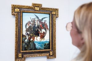 A blonde woman with glasses looking at a framed painting on a wall.