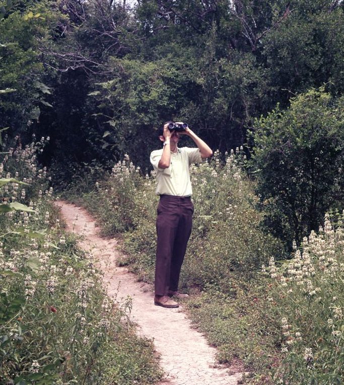 A vintage photo of a man in slacks holding binoculars and looking toward the sky surrounded by a forest