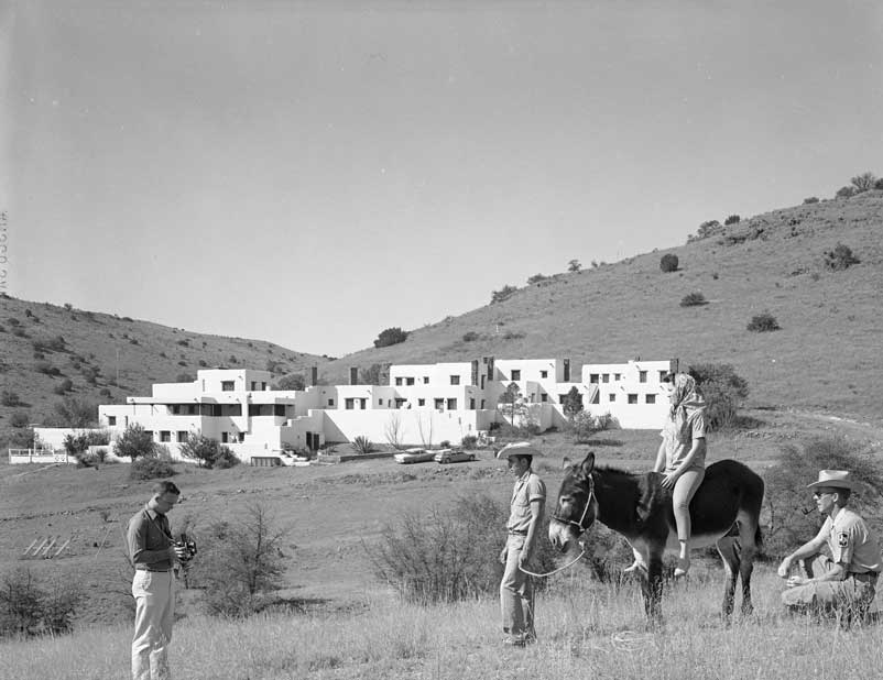 A man taking a picture of two men next to a woman on a mule in front of a white adobe lodge