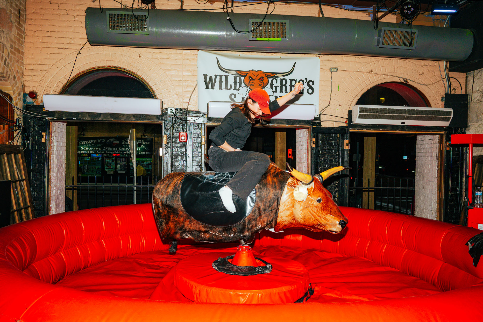 A woman in a red cap riding a mechanical bull in a red pit.