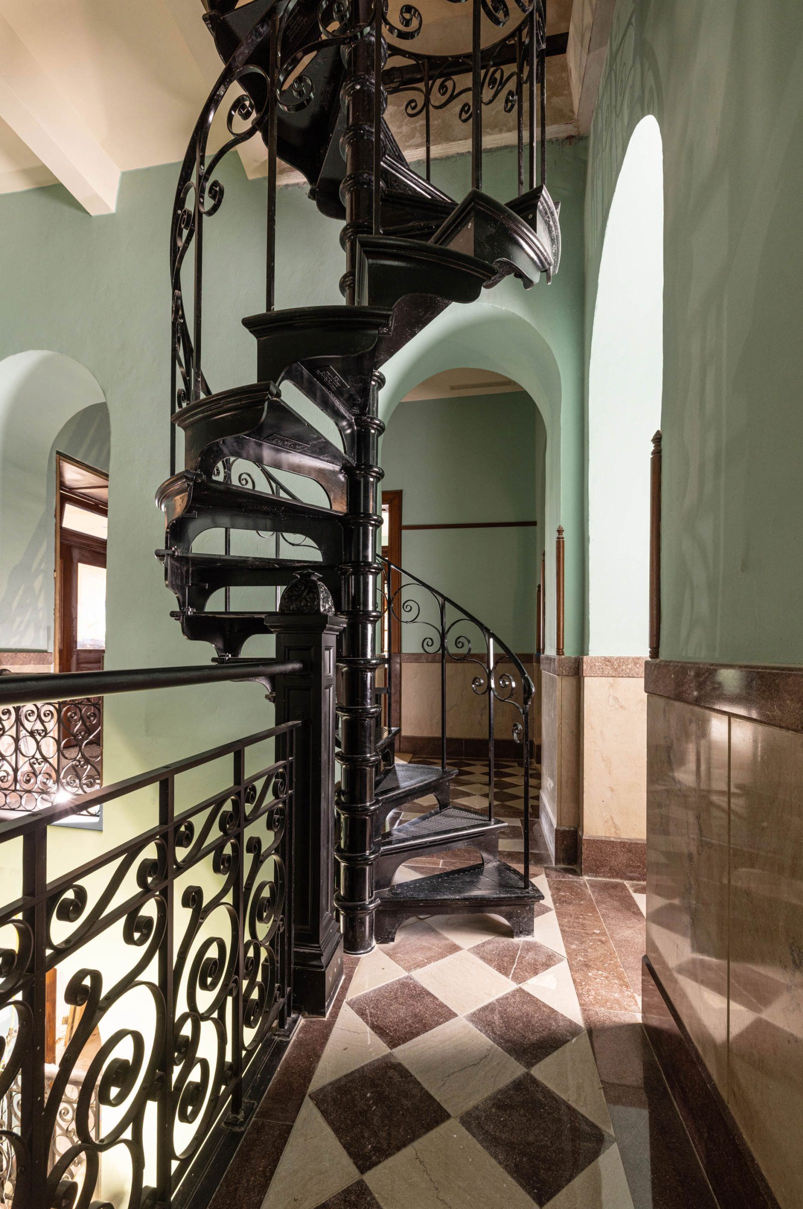 A black spiral staircase inside a hall painted a light seafoam with a black and white harlequin patterned floor