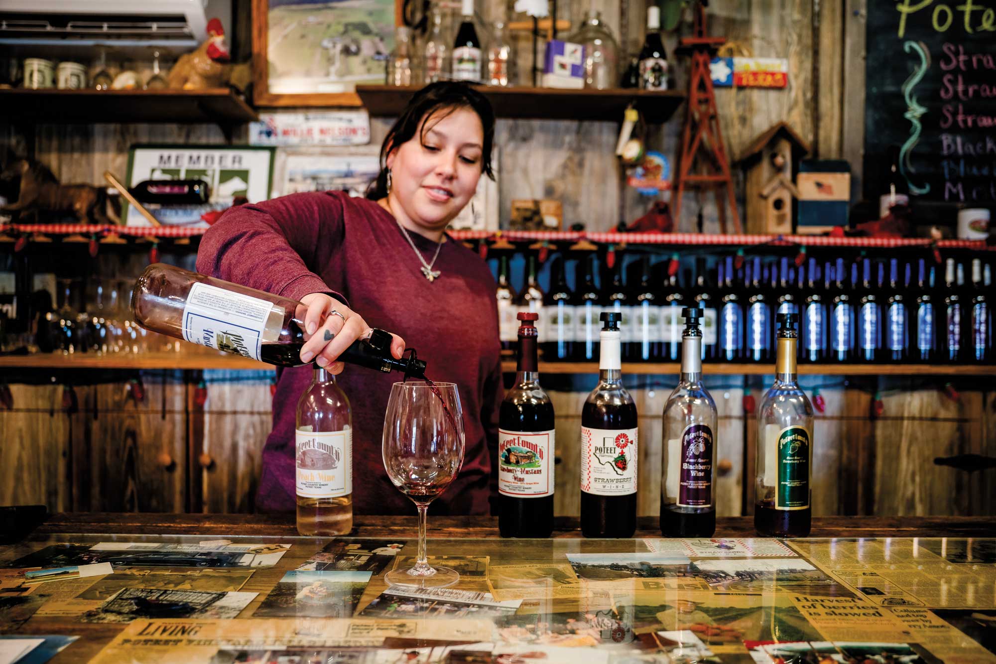 A woman in a burgundy shirt and necklace pouring red wine into a wine glass atop a bar with numerous wine bottles next to and behind her