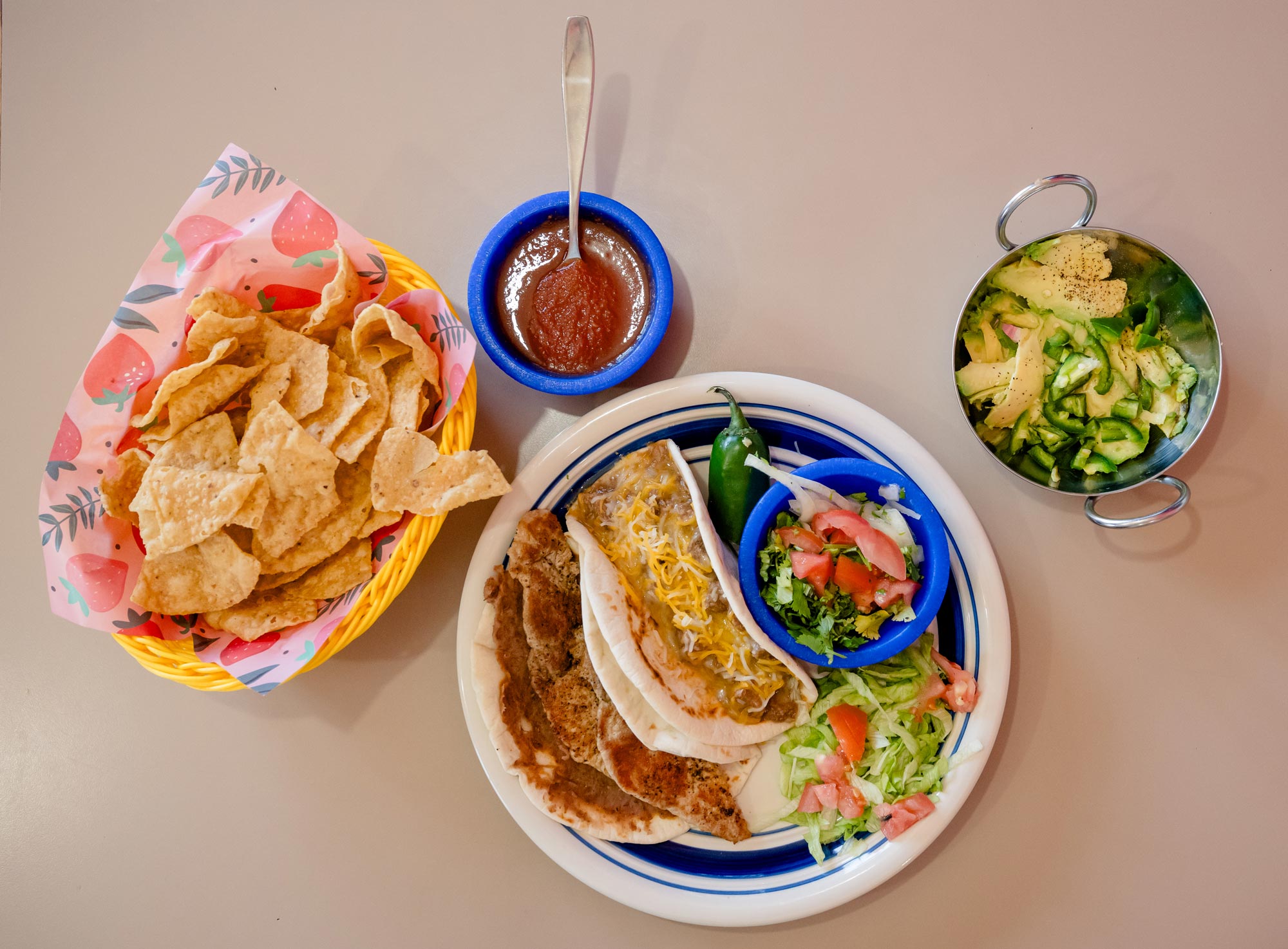 A basket of tortilla chips, a blue cup of red salsa with a spoon in it, a metal bowl with avocado and green peppers, and a white-and-blue plate of tacos, a jalapeño, lettuce, onions, and cilantro