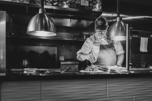 A chef wearing a baseball cap seasoning a dish under lamps