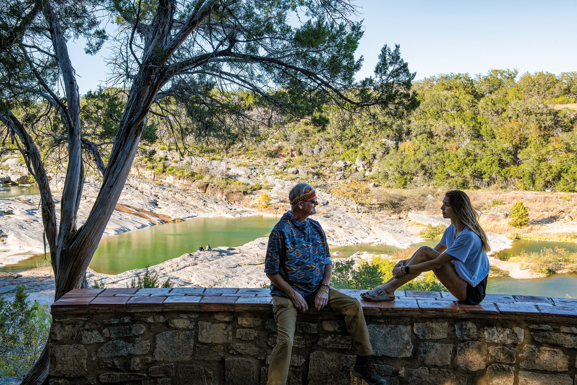 Two people sit on a ledge, a man with his legs on the ground and a woman with her legs on the ledge, with a tree next to them