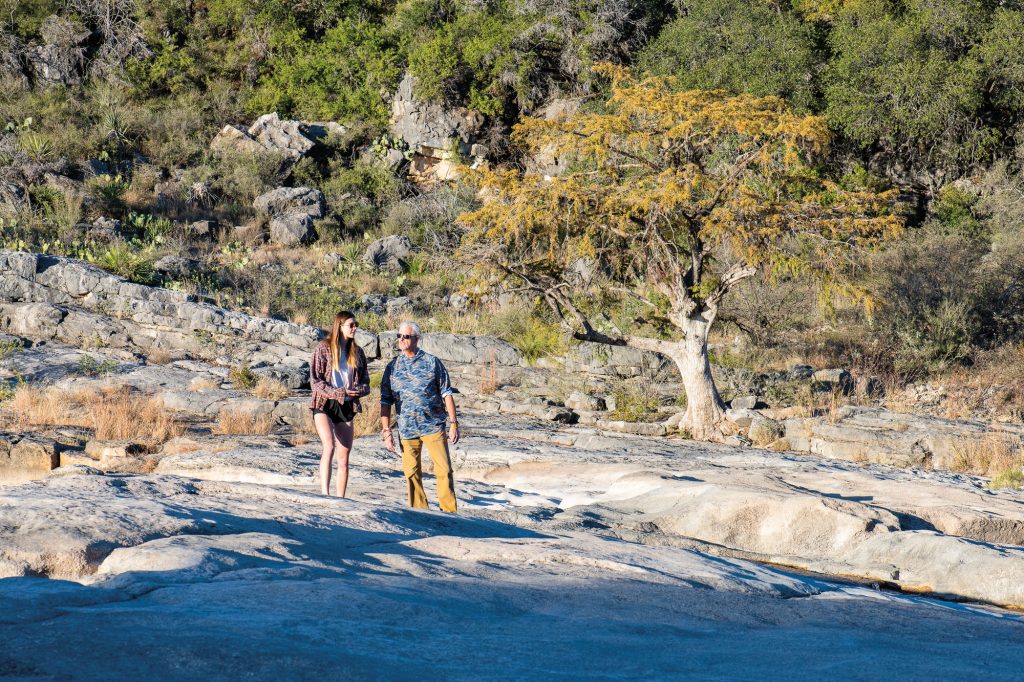 Two people walking on a craggy area, with a tree and a rock formation behind them