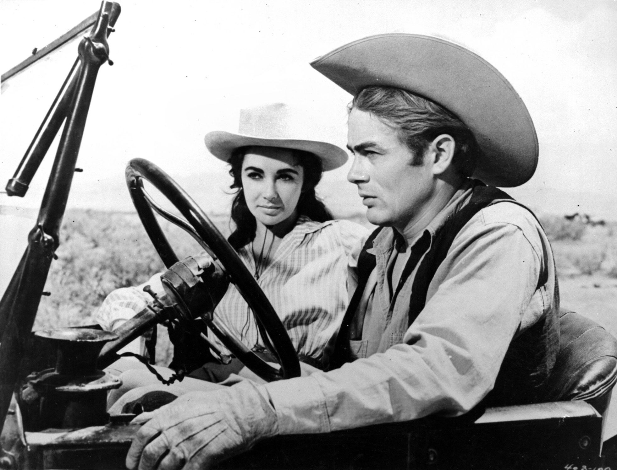 A black and white photo of a man and woman sitting in an old car in a field while wearing cowboy hats