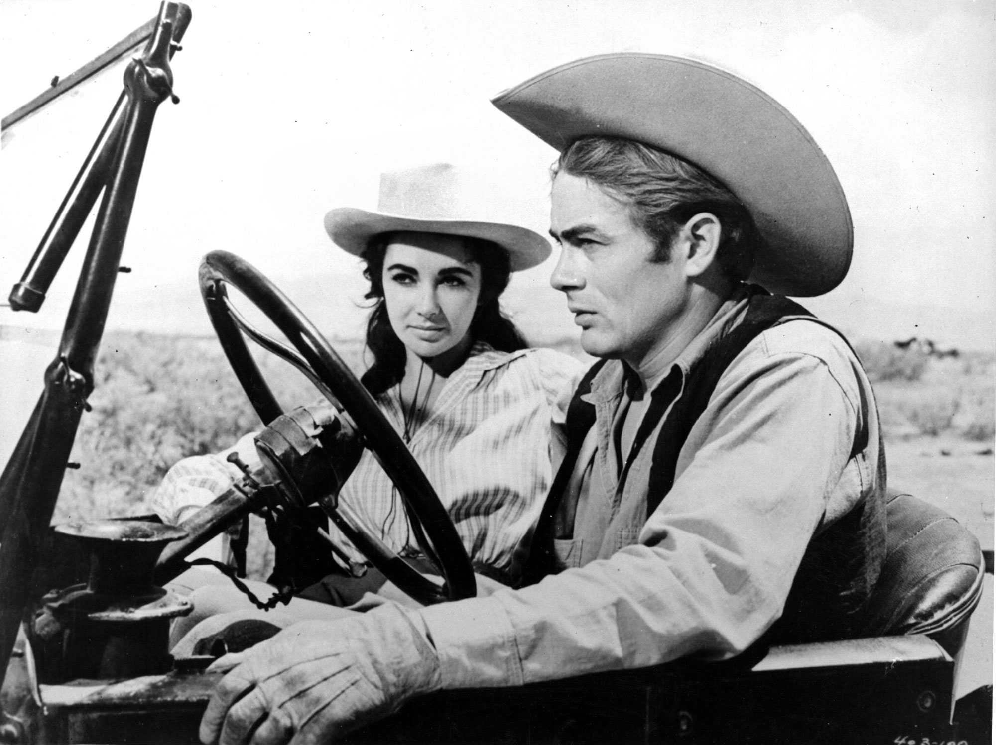 A black and white photo of a man and woman sitting in an old car in a field while wearing cowboy hats