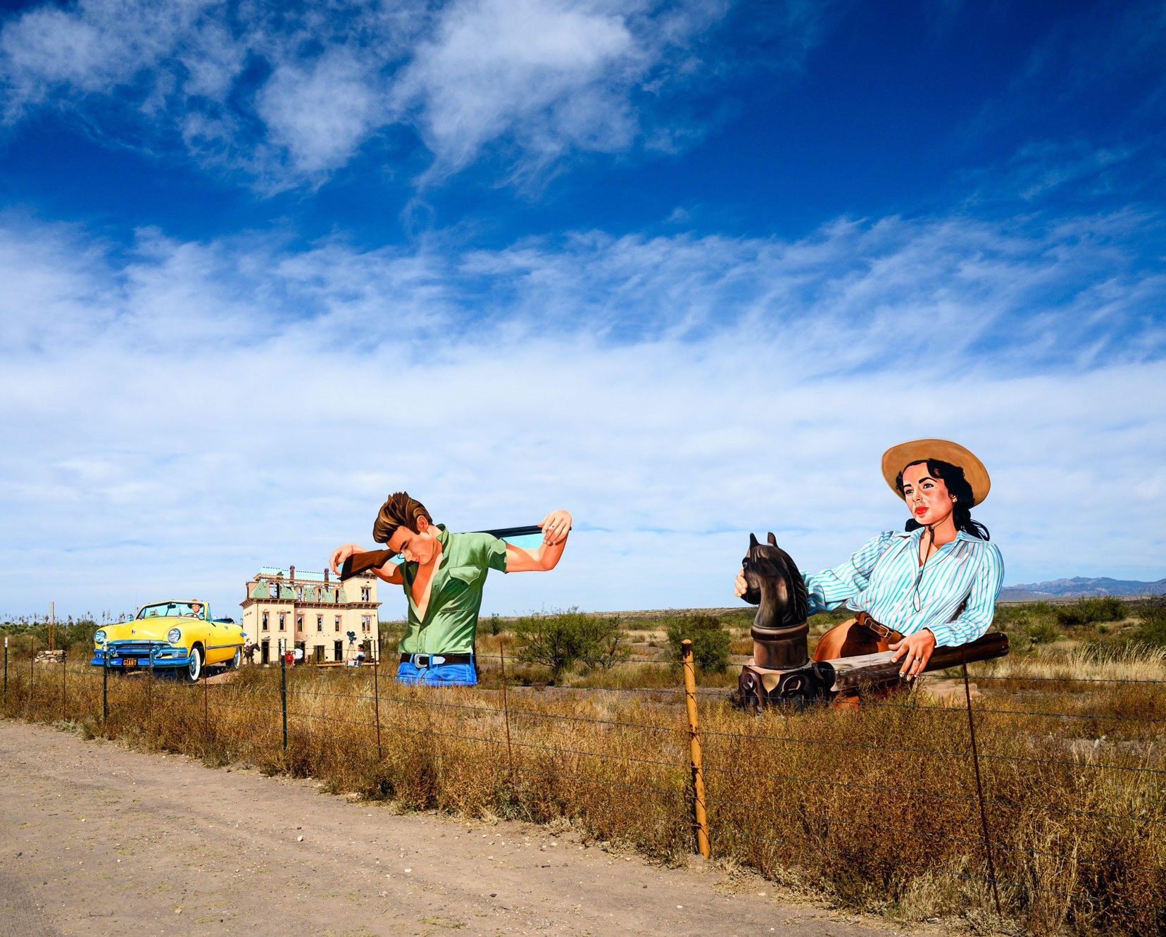 Several large painted wooden cutouts of a woman leaning on a fence, a man with a shotgun, a large mansion, and a retro yellow car stand in a field beside a road.