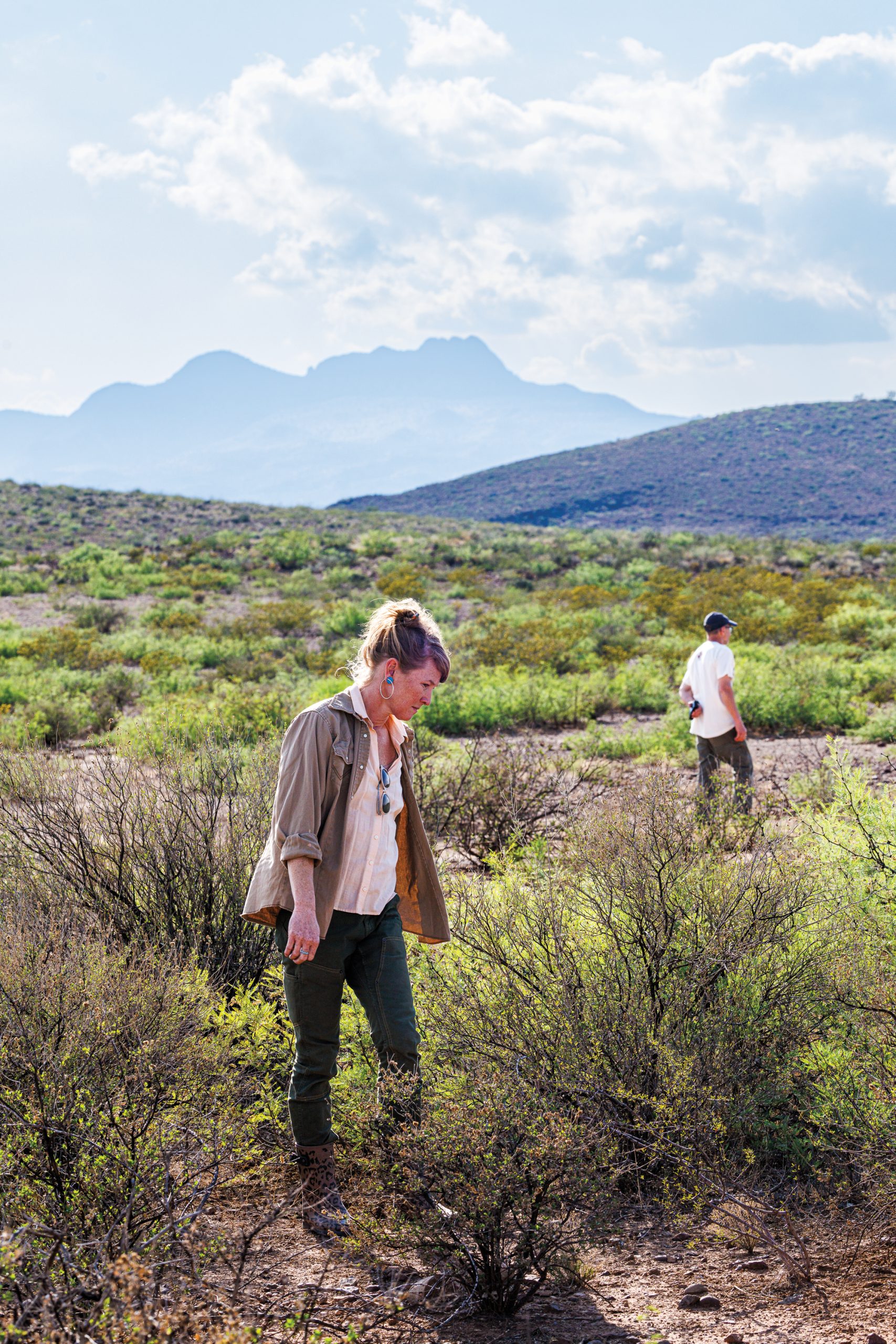 A man and woman stand in a shrubby desert landscape with a mountain in the background