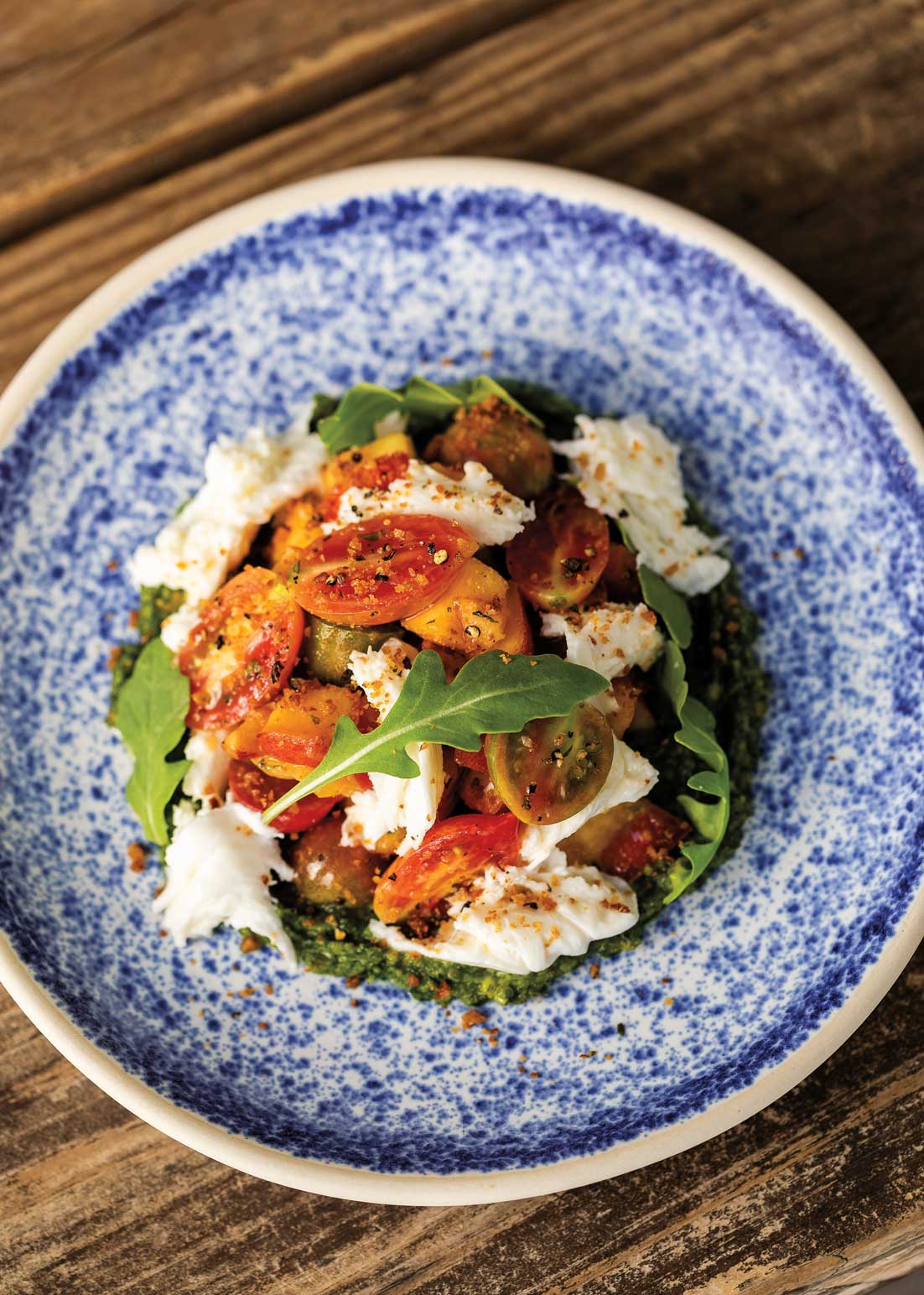 A white bowl with blue dots filled in the middle with arugula, mozzarella, a nectarines covered in seasoning