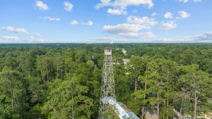 A large, rusted tower in the middle of a lush, green forest under blue skies filled with clouds