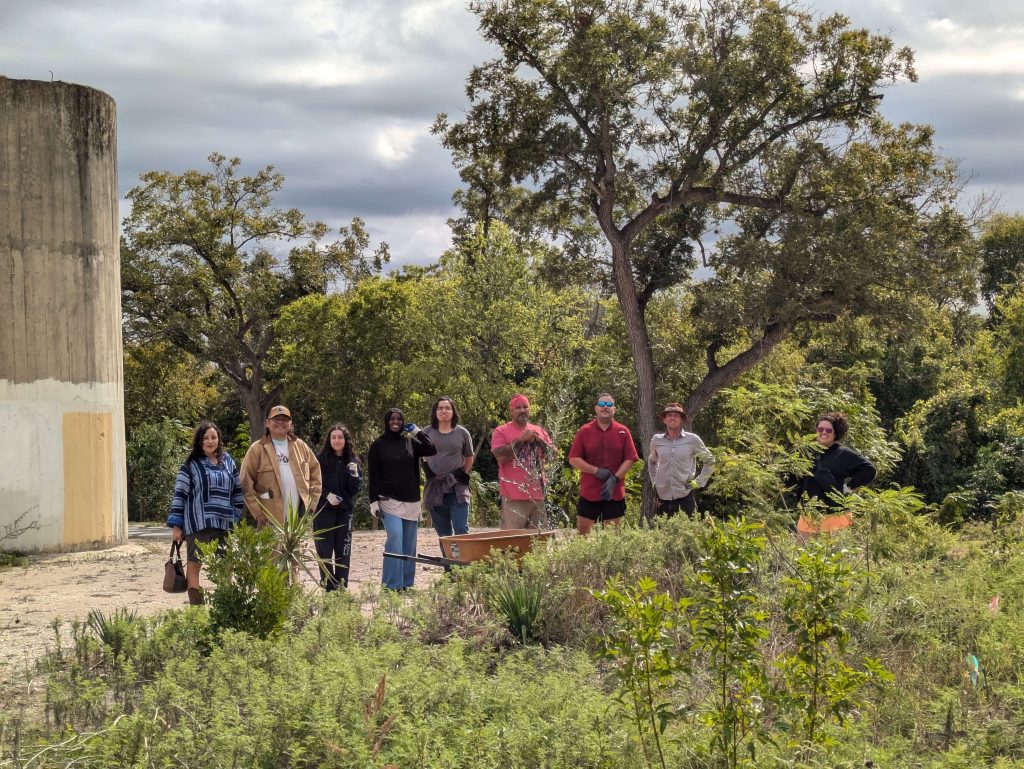 Several people dressed in gardening clothes stand in a lush outdoor area in front of a wheelbarrow