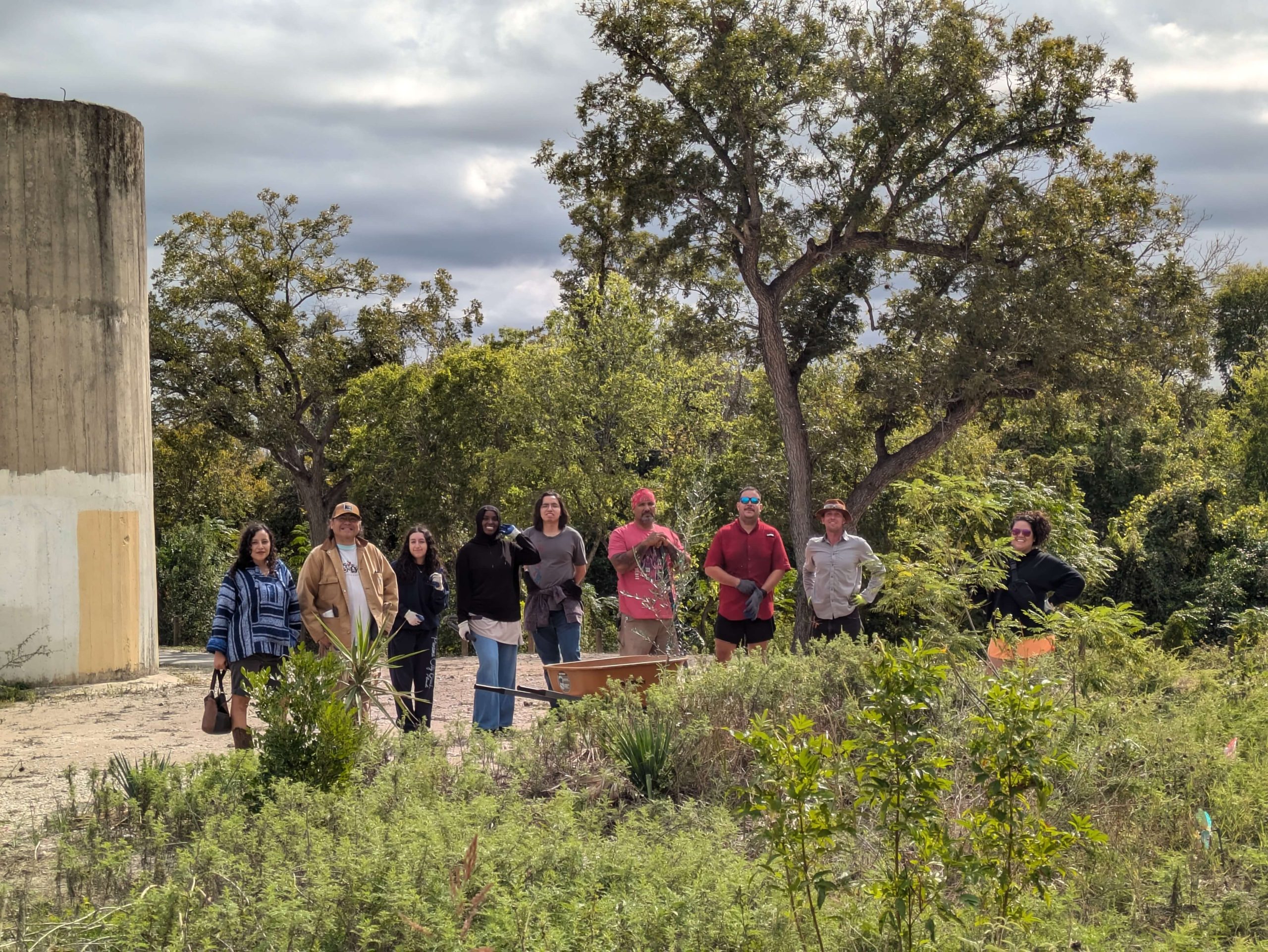 Several people dressed in gardening clothes stand in a lush outdoor area in front of a wheelbarrow