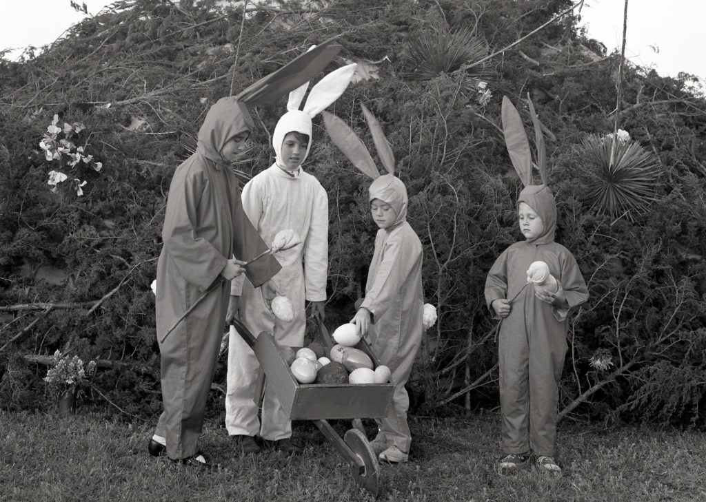 Four children in Easter Bunny costumes surround a wheelbarrow filled with plastic eggs, behind them is a large pile of brush with a wooden cross atop it