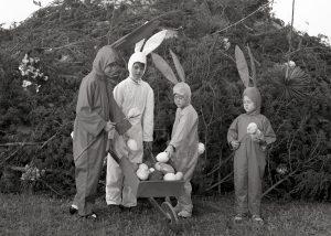 Four children in Easter Bunny costumes surround a wheelbarrow filled with plastic eggs, behind them is a large pile of brush with a wooden cross atop it