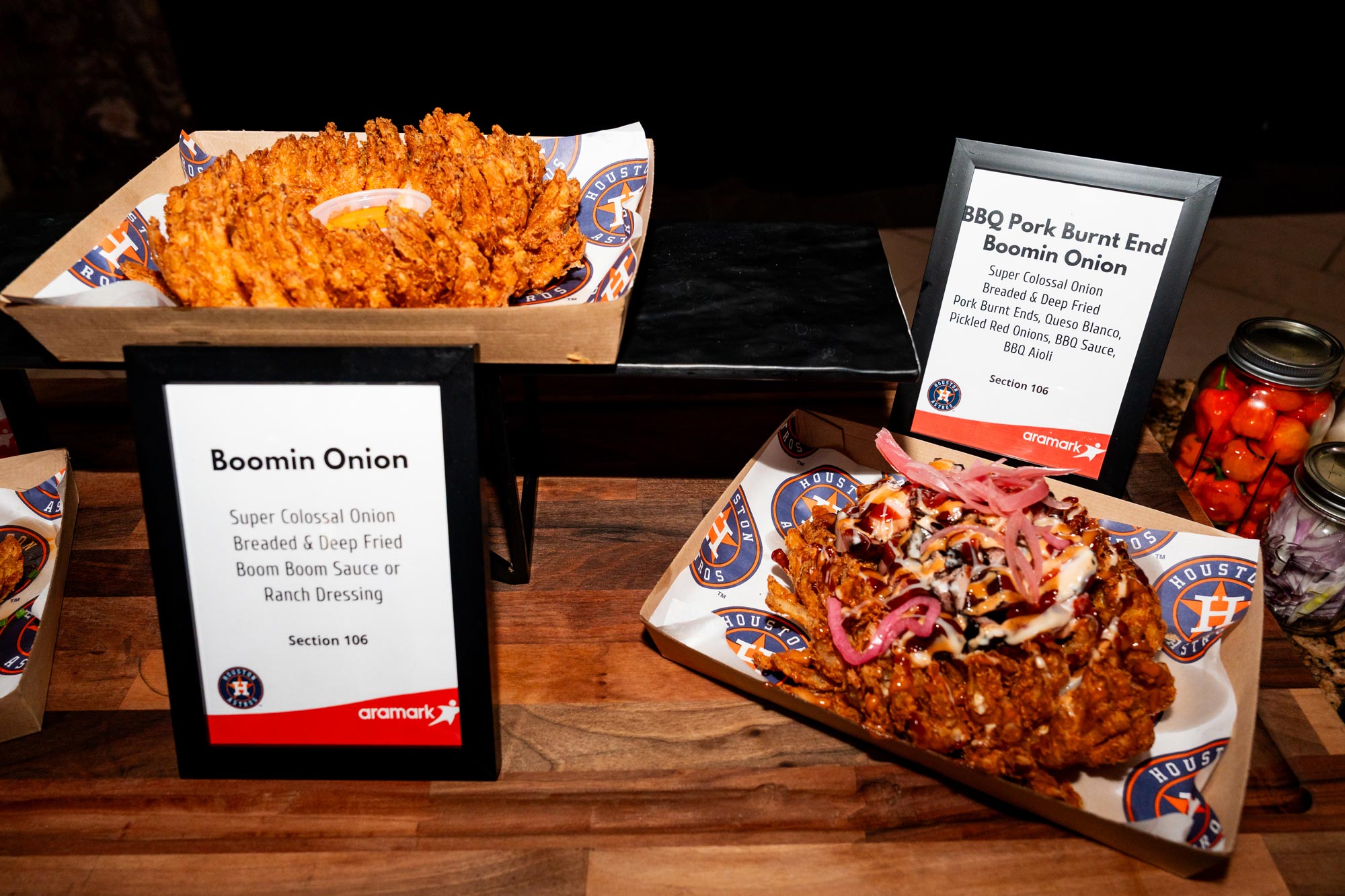 A large fried onion with a plastic cup of yellow sauce in the middle on a Houston Astros tray above a barbecue pork burnt end fried onion with pickled red onions and queso on top in a similar Houston Astros container.