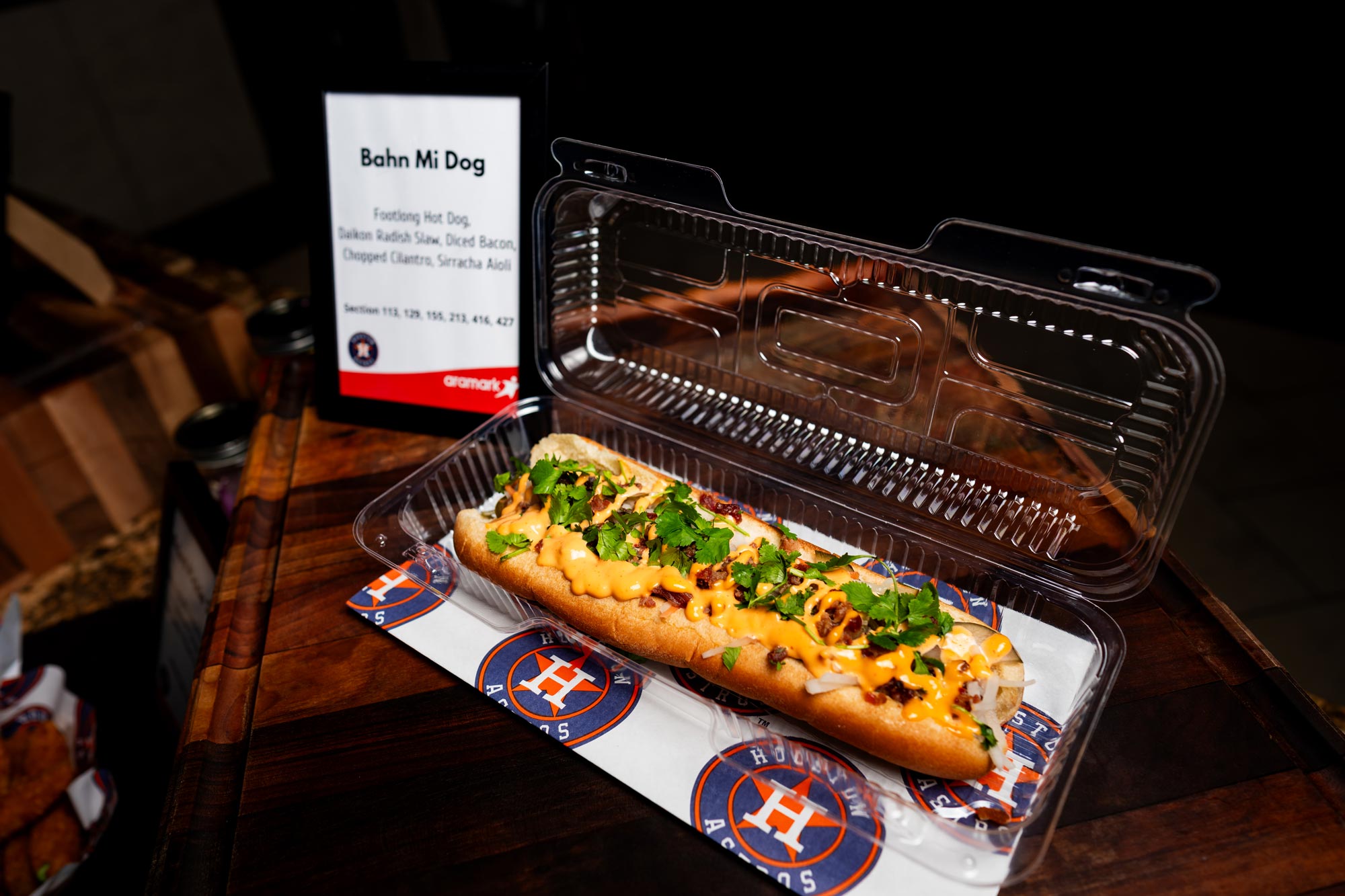 A hot dog covered in cole slaw, bacon, and cilantro in a plastic container with a Houston Astros paper beneath it