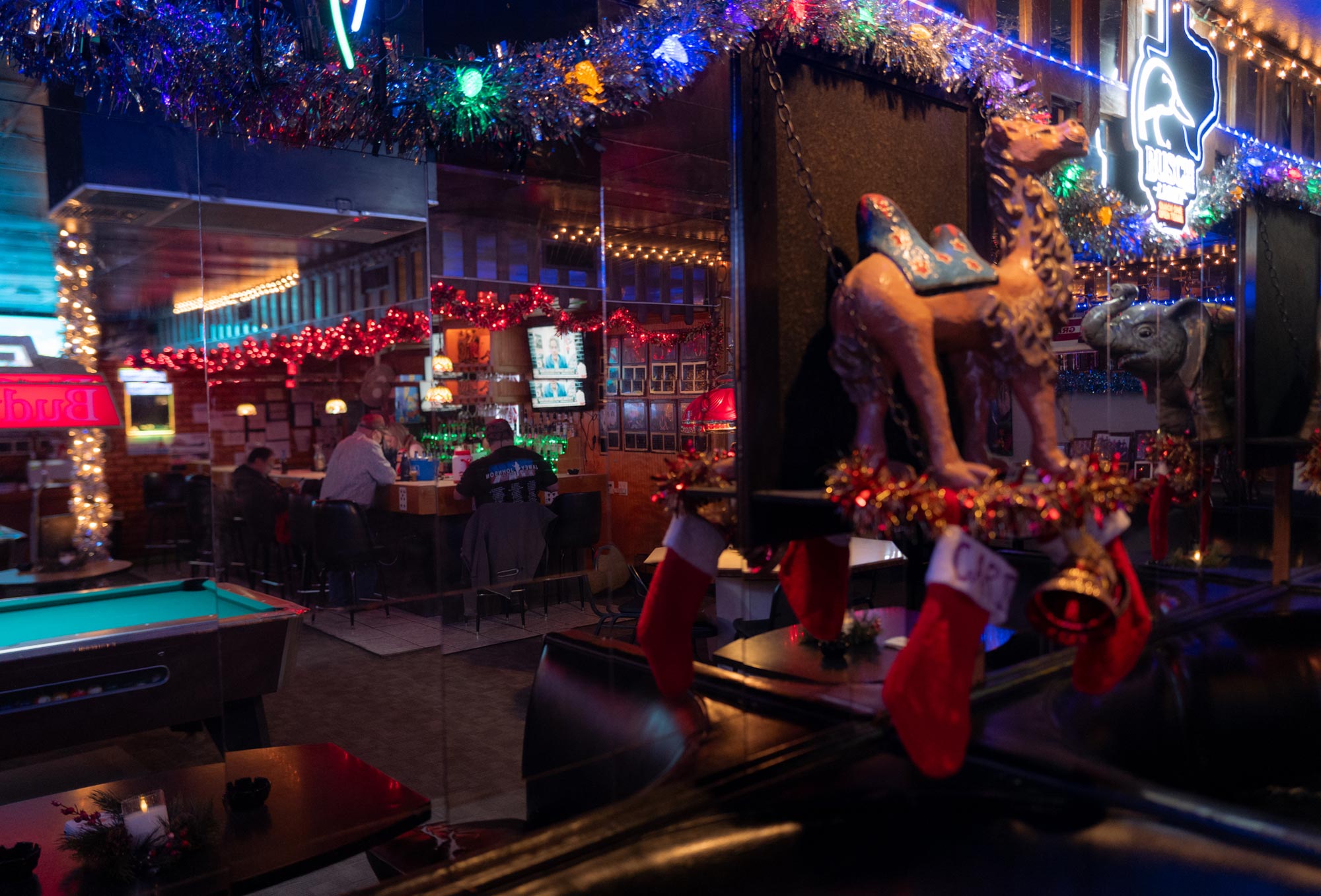 An image of the inside of a bar, with a tiled mirror reflection showing three patrons sitting at the bar, plus hanging Christmas stockings and statues of a camel and an elephant.