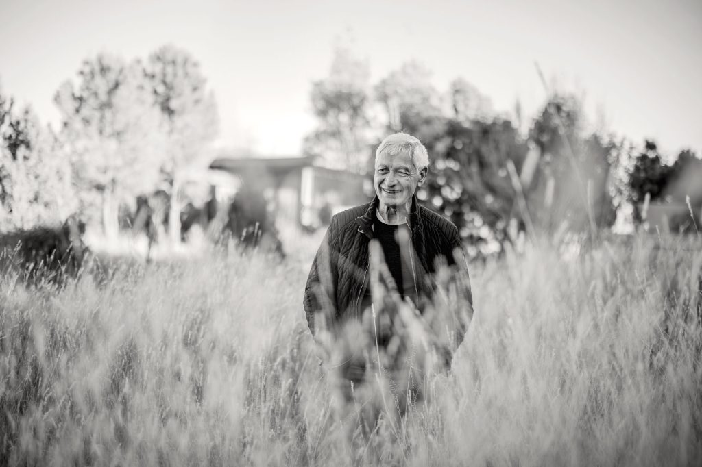 A black-and-white photo of a smiling man with white hair, partially obscured by tall grass in the foreground