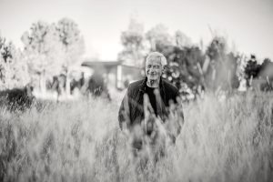 A black-and-white photo of a smiling man with white hair, partially obscured by tall grass in the foreground