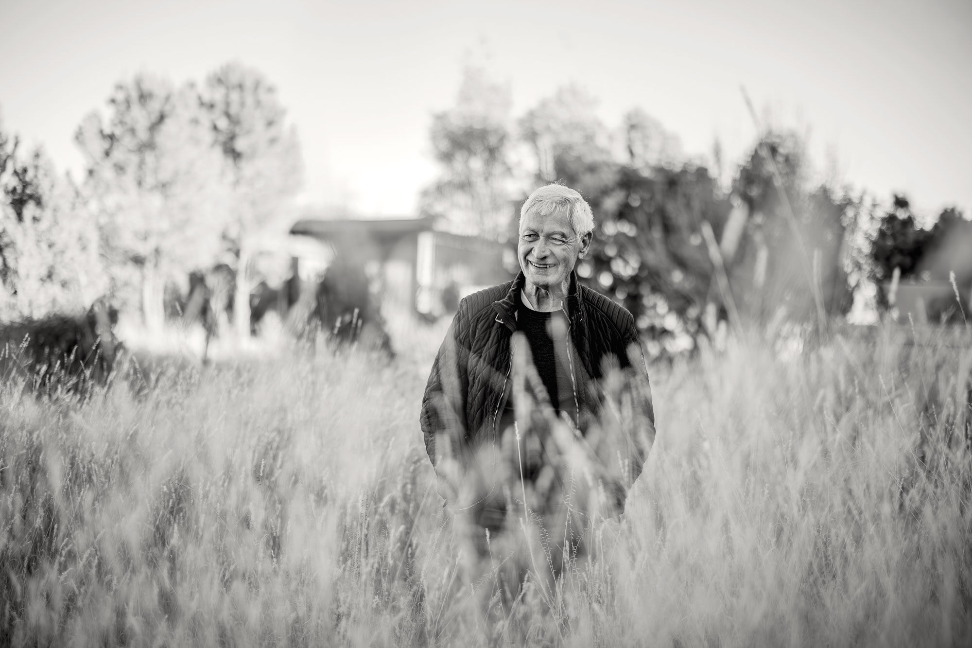 A black-and-white photo of a smiling man with white hair, partially obscured by tall grass in the foreground
